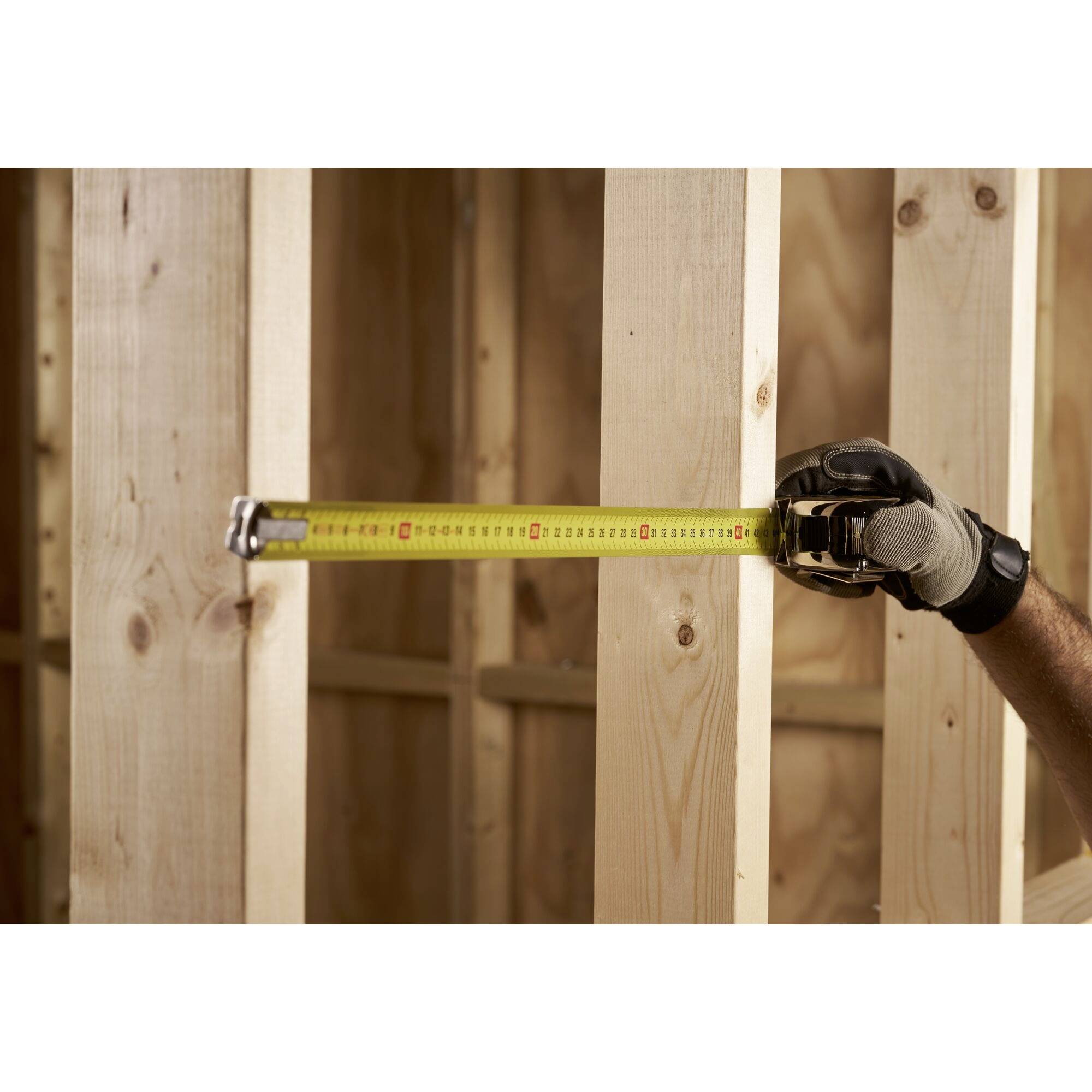 A worker is measuring the distance between vertical wooden posts in an unfinished frame construction.