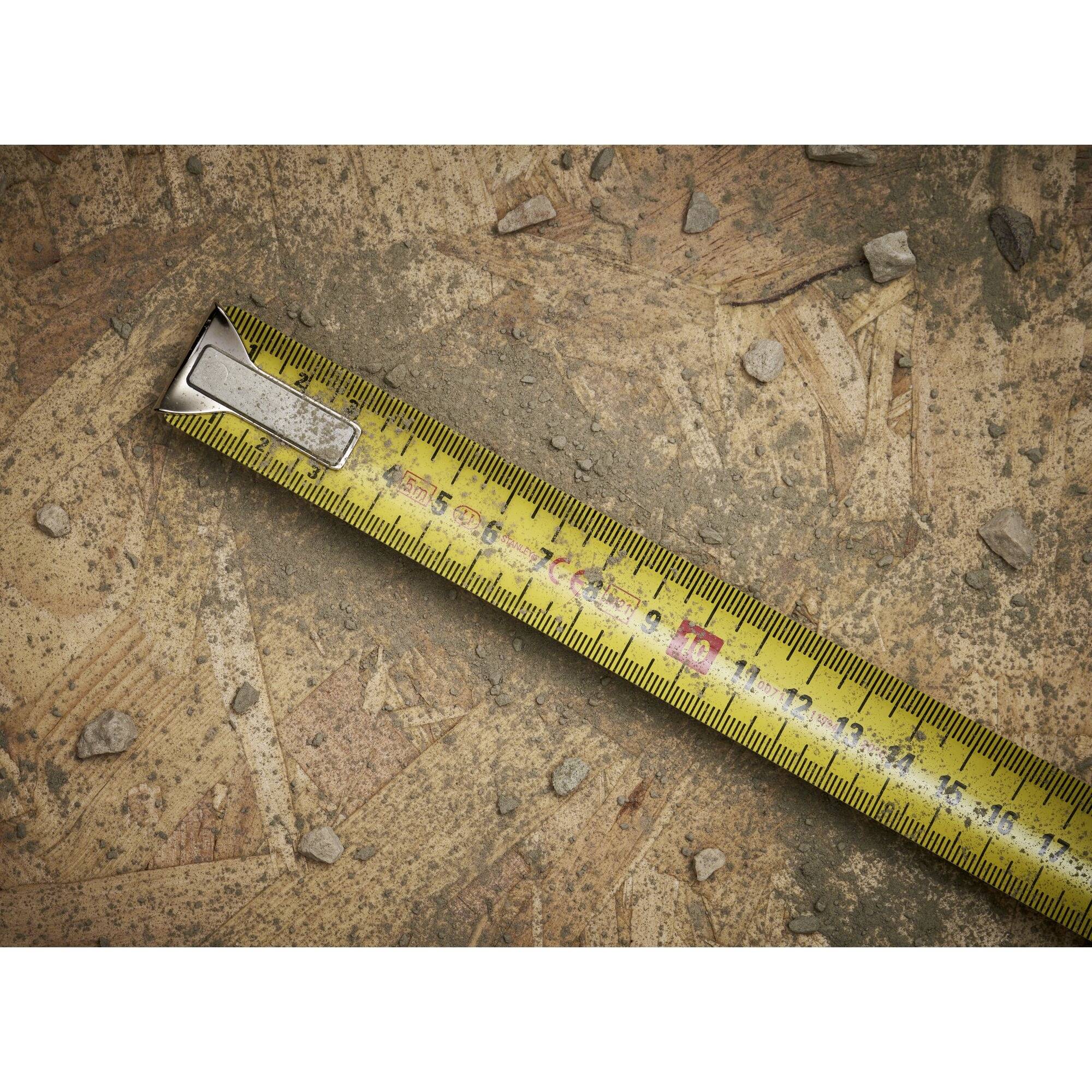 A yellow tape measure lies slightly diagonally on a rough, stony wooden surface. It indicates a length of approximately 17 cm.