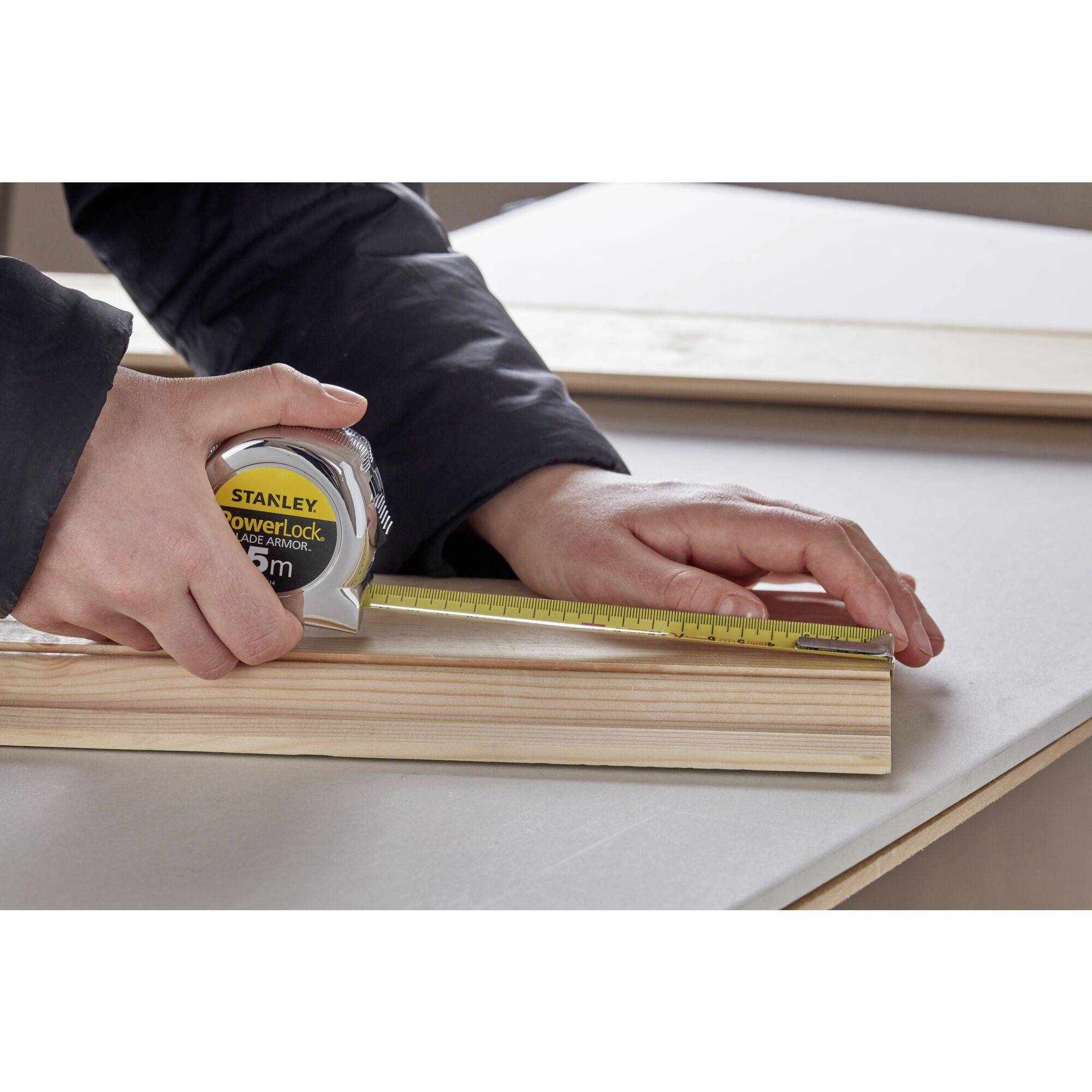 A person is measuring a wooden board with a yellow tape measure on a table.