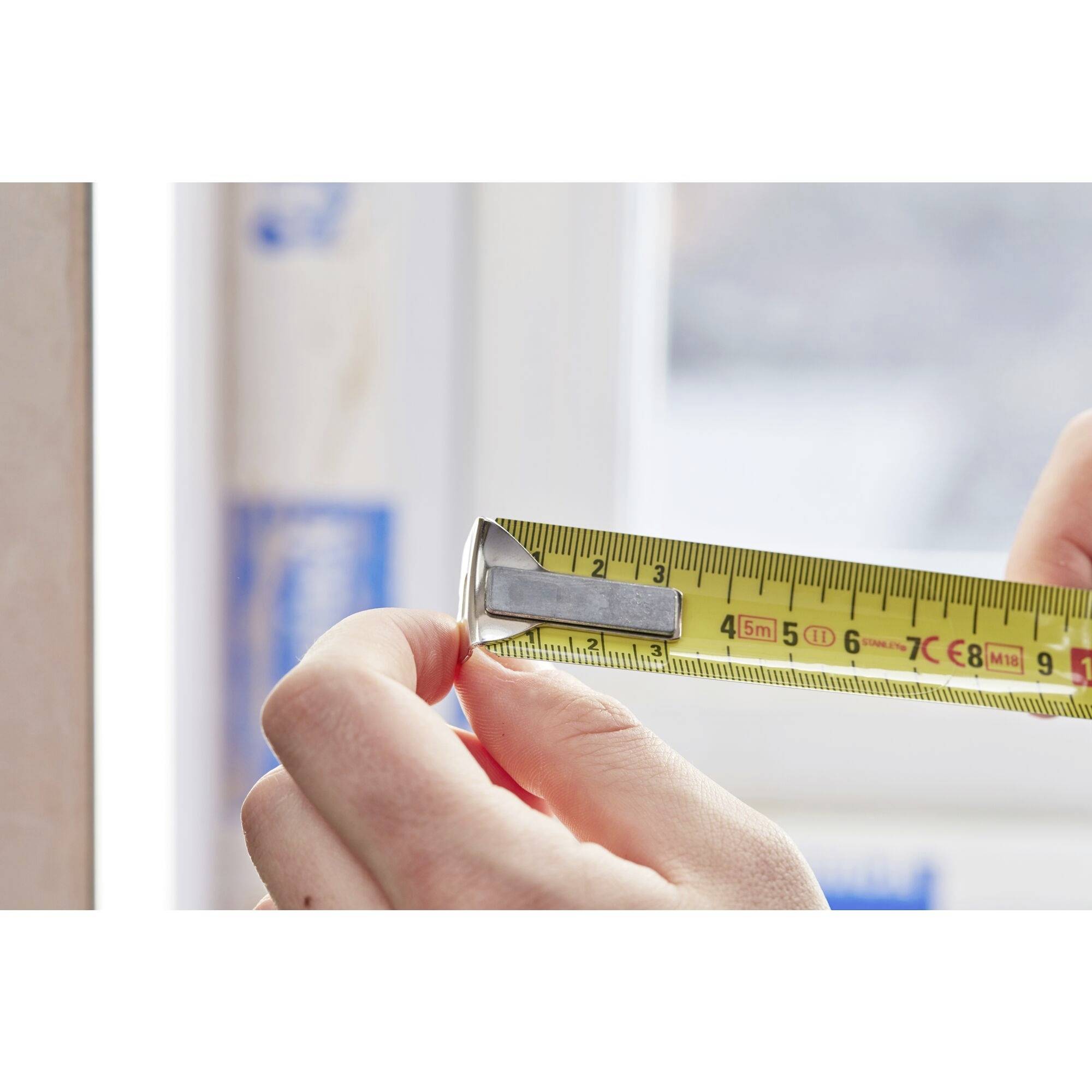A hand is holding a yellow tape measure against a window frame. The tape measure shows approximately 7.5 centimetres.