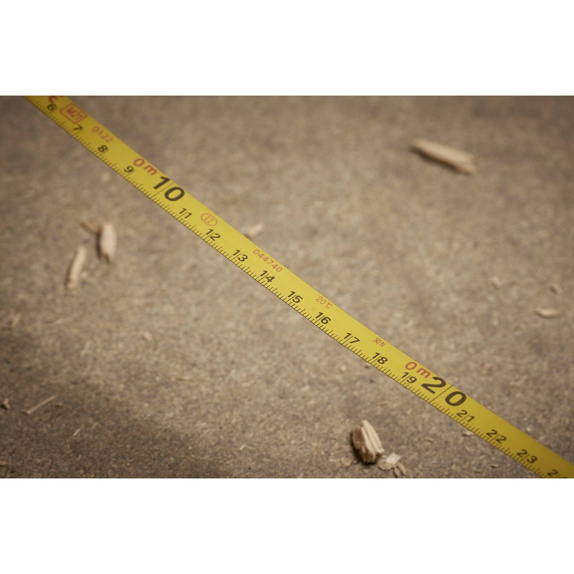 A yellow tape measure lies diagonally on a sandy ground. It shows centimetre markings from 0 to 20. Wood chippings are visible in the background.