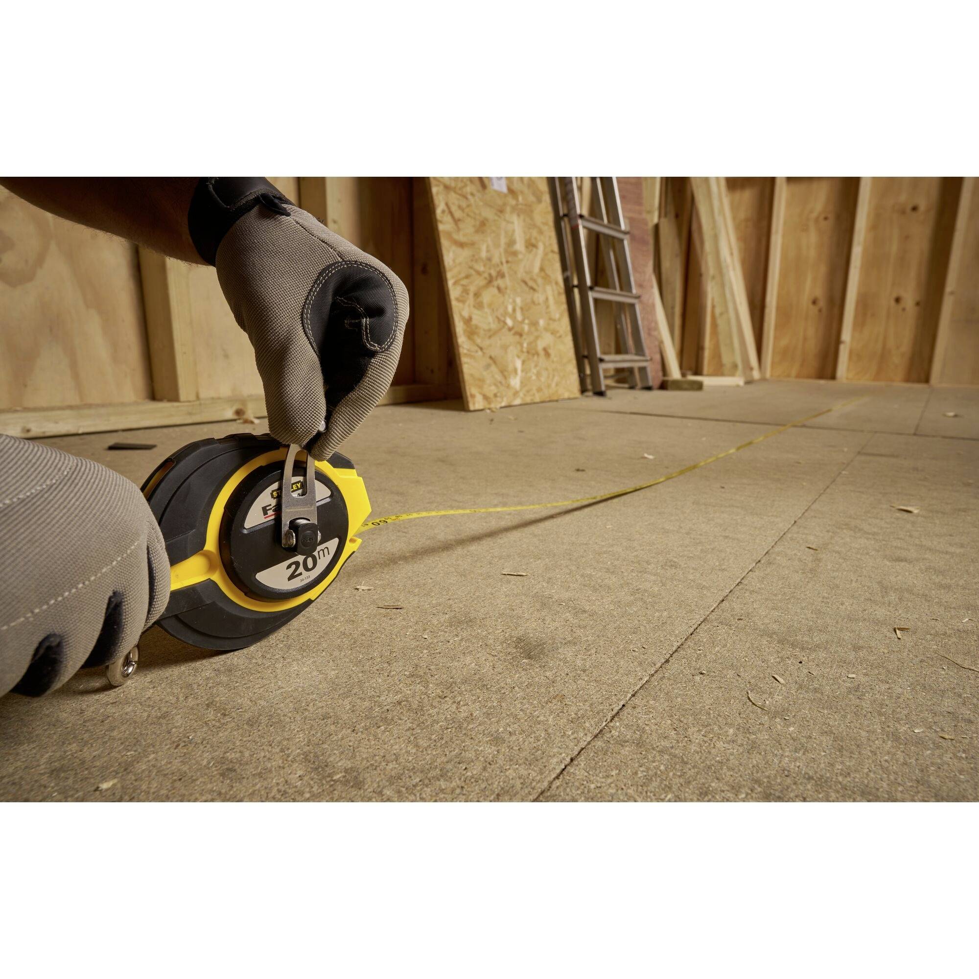 A gloved hand measuring with a tape measure on a wooden floor in an unfinished room with wooden panelling and a ladder.