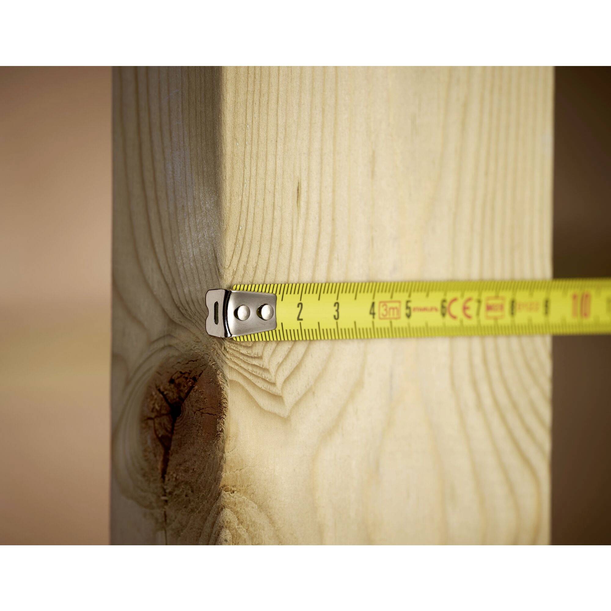 A tape measure is measuring the diameter of a wooden post. The tape shows approximately 10 cm. The post has a visible wood grain and a knot hole.