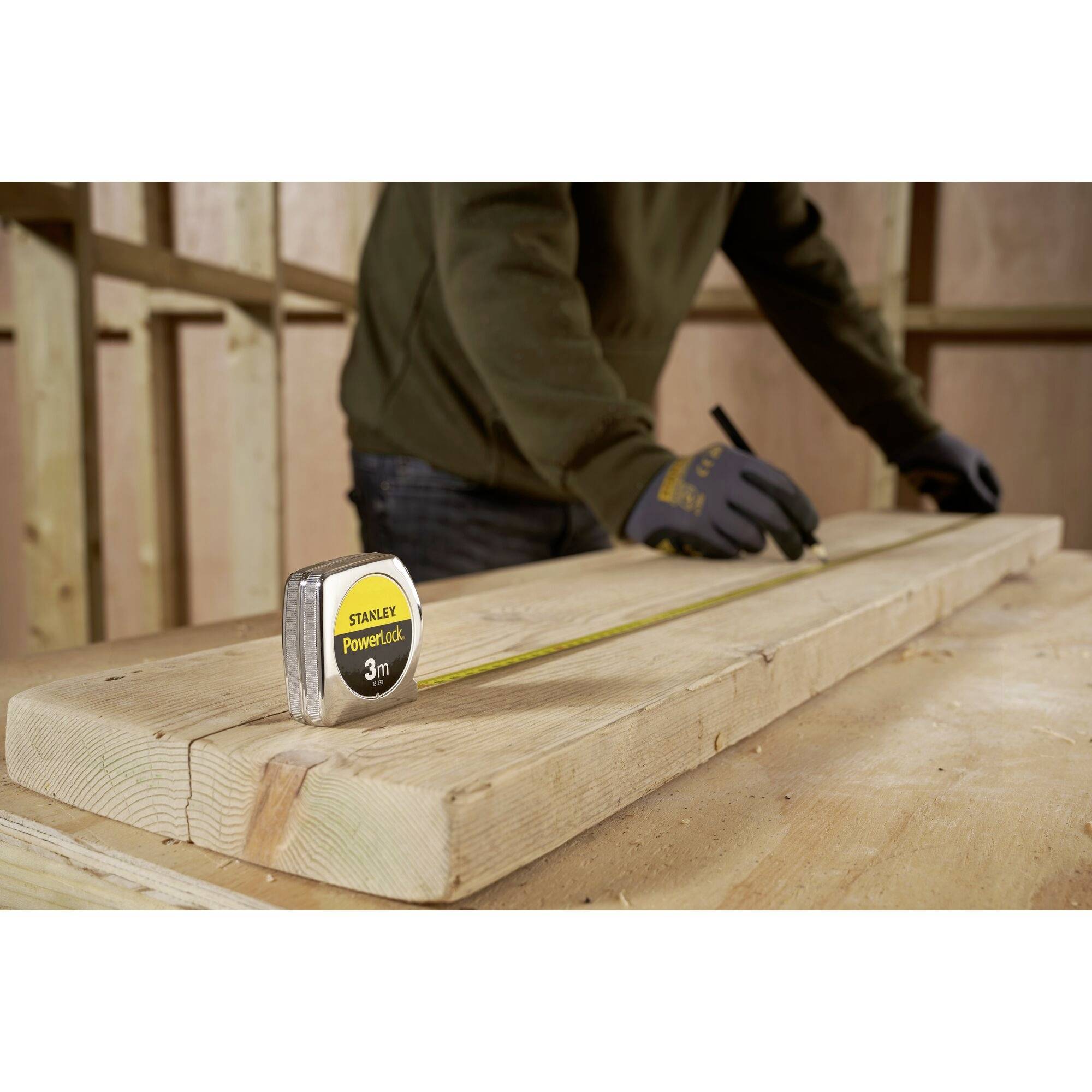 A person wearing gloves is measuring a wooden board with a 3-metre measuring tape on a construction site.