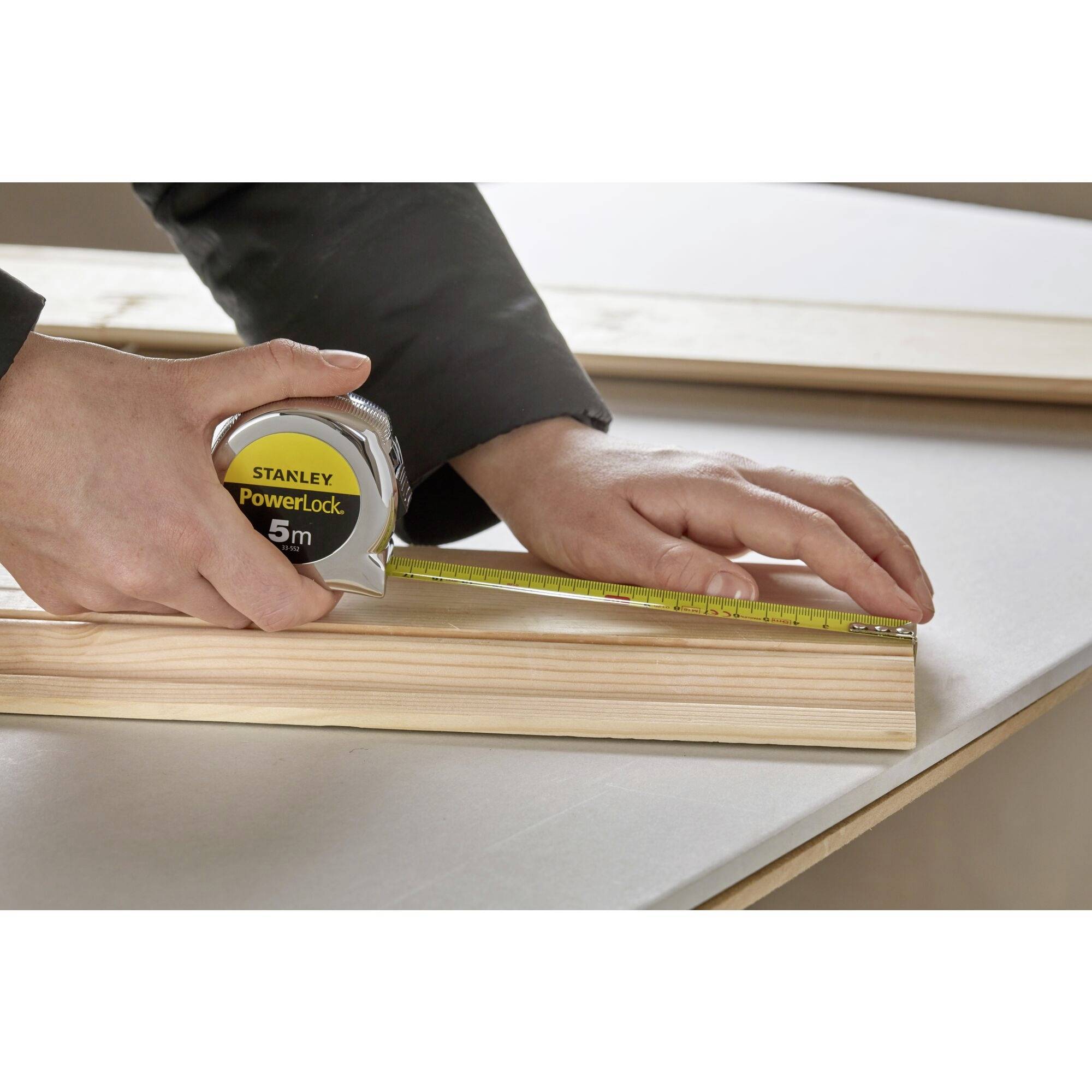 A hand holds a tape measure running along a piece of wood. The tape measure shows 5 metres. Gloves protect the hands.