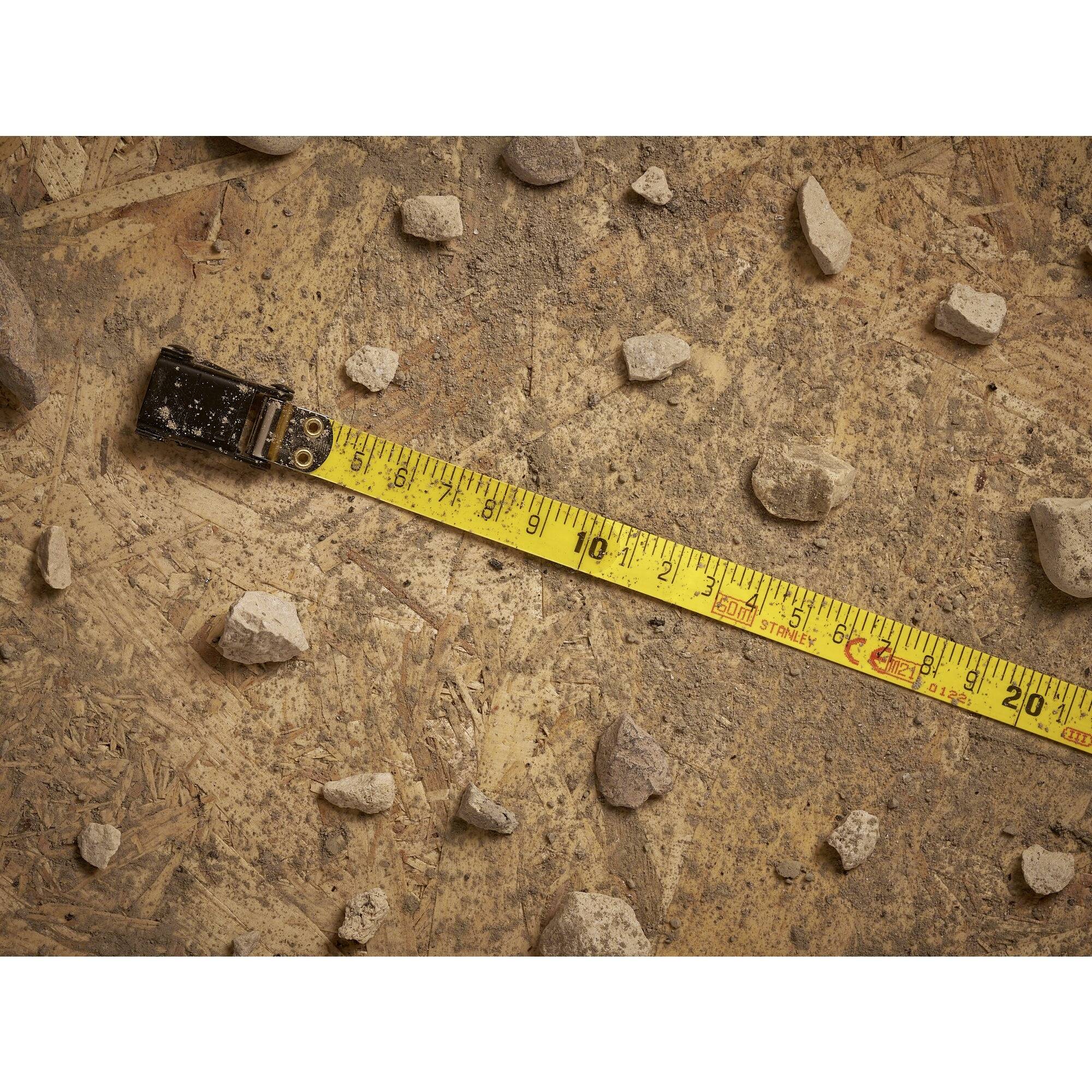 A yellow tape measure lies diagonally on a rustic wooden floor, surrounded by scattered small stones.