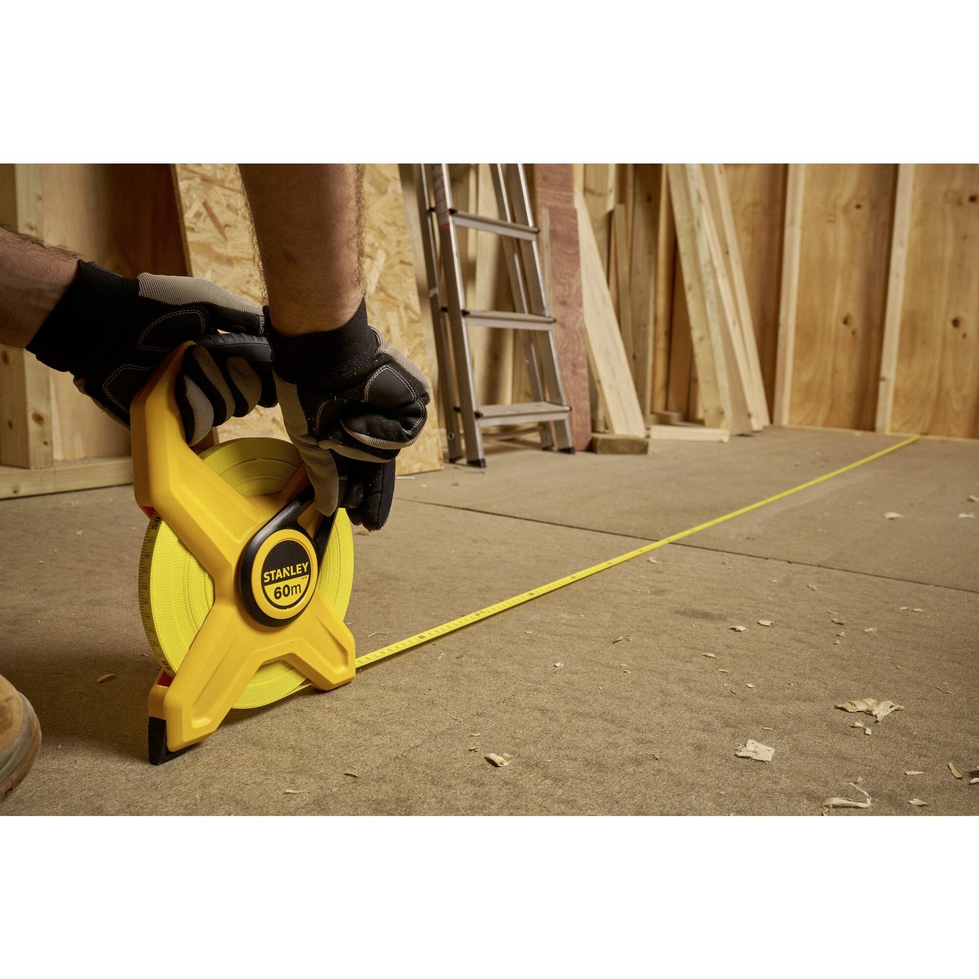 A person wearing work gloves is measuring with a yellow tape measure on a wooden workbench in a workshop. Wooden materials are visible in the background.