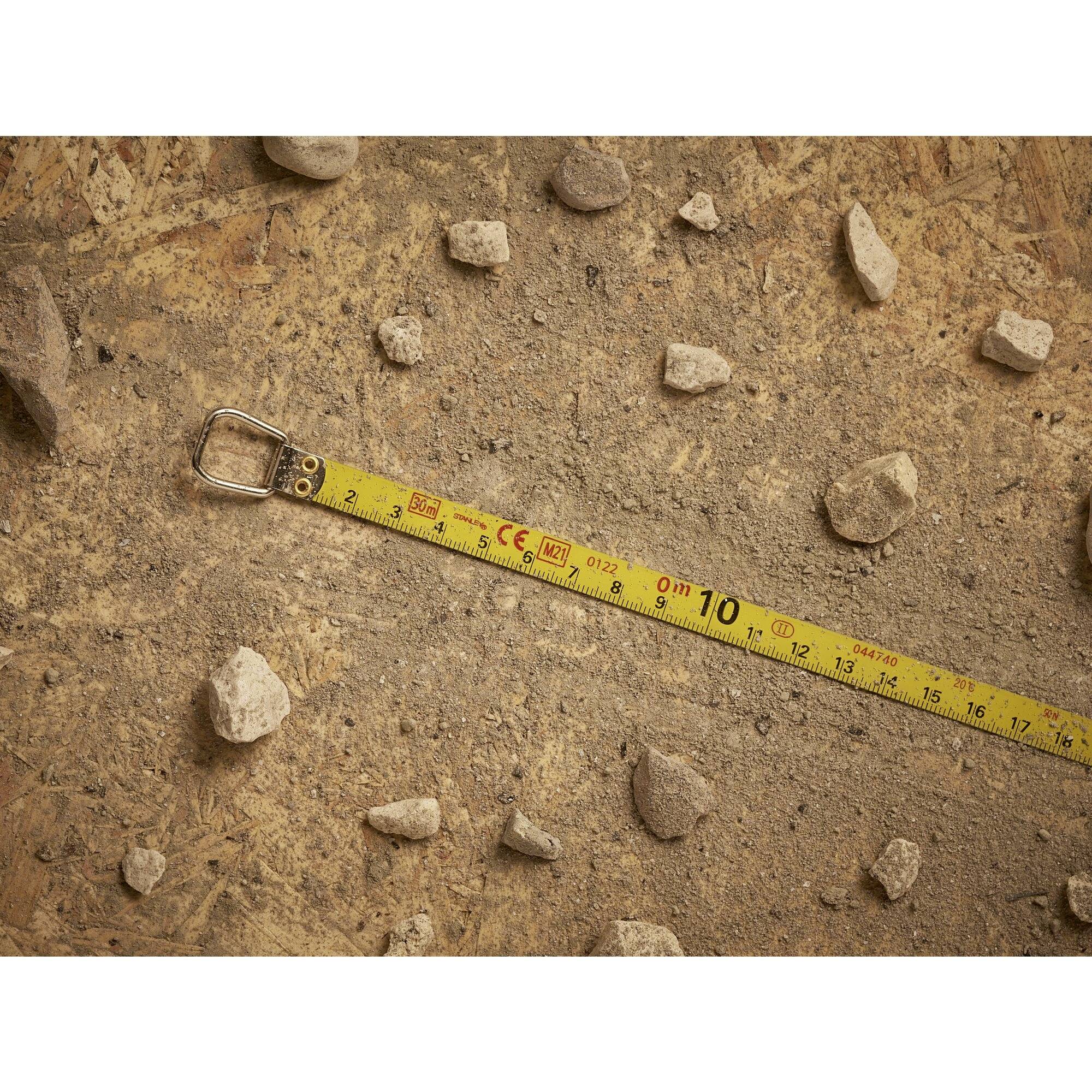 A yellow tape measure lies on an earthy ground with scattered small stones. The tape shows centimetre and metre measurements.
