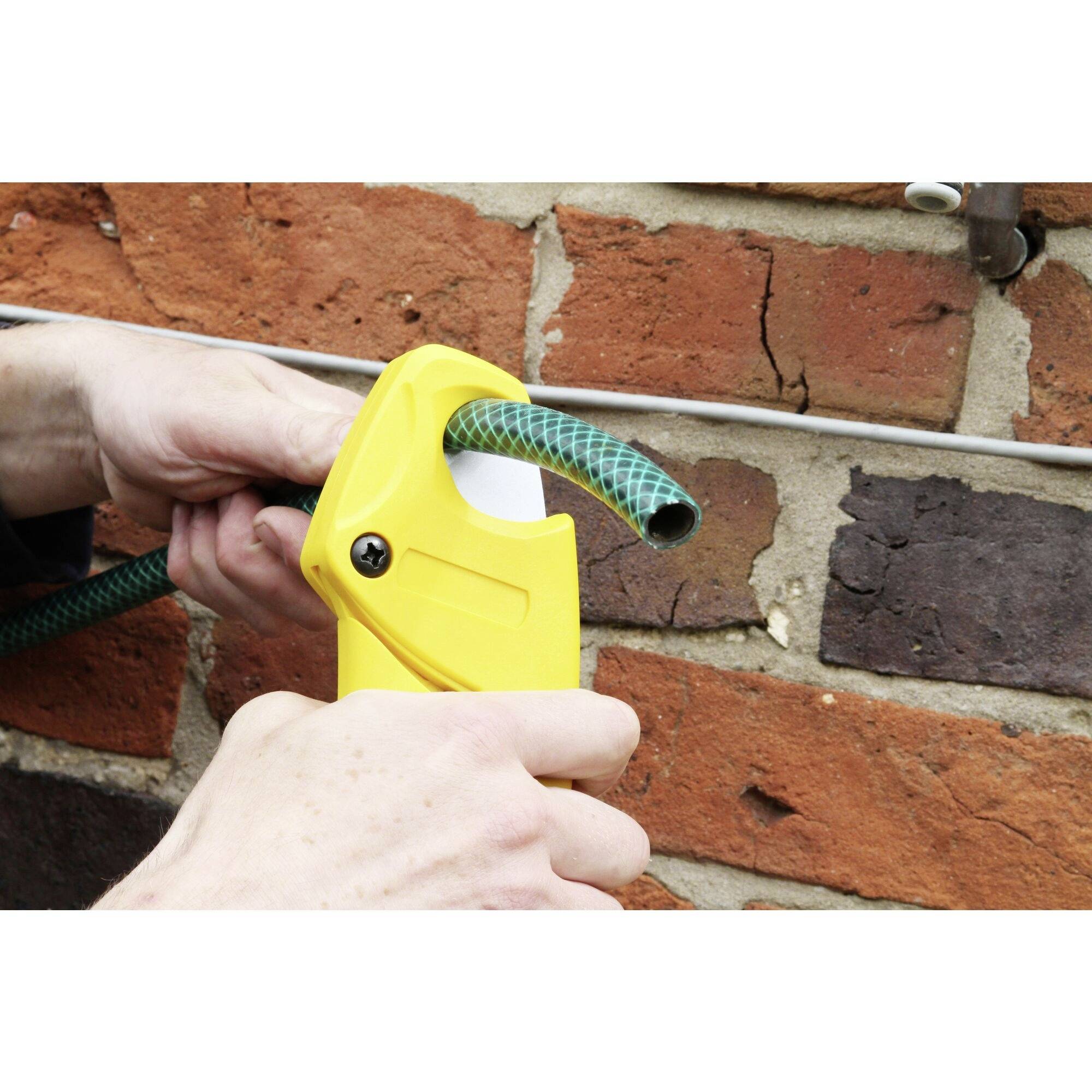 A person is cutting a green garden hose with yellow scissors in front of a brick wall.