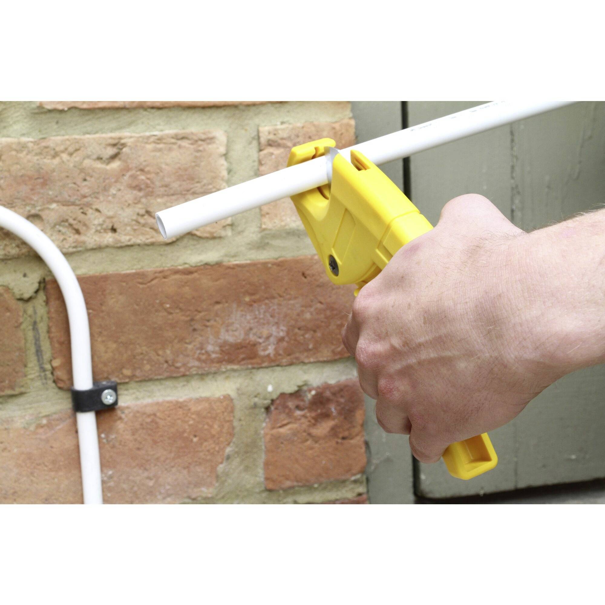 A hand is holding a yellow pipe cutter and cutting a white pipe in front of a brick wall.