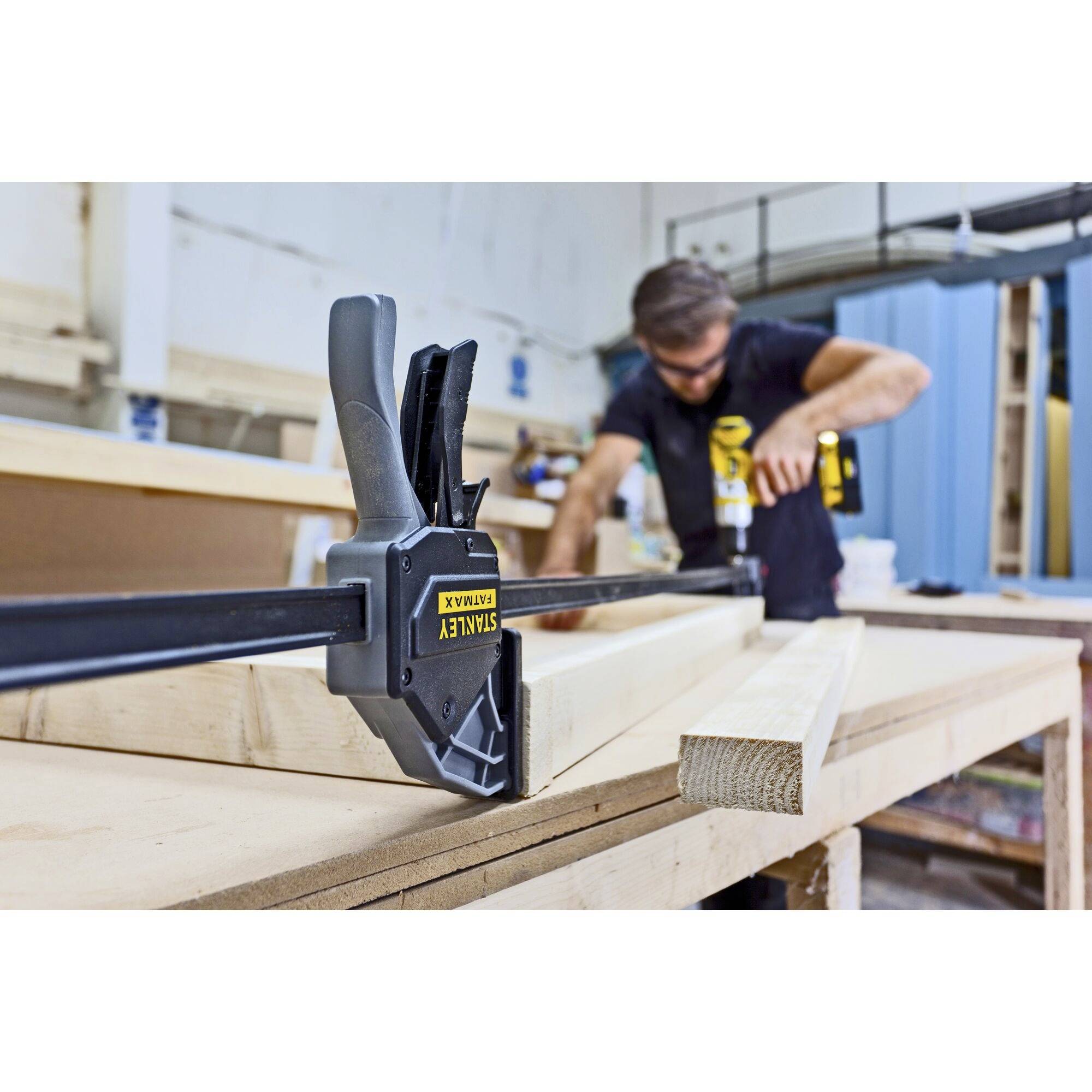 A man is working in a workshop, using a clamp to secure wooden pieces on a table. He is wearing safety glasses and a tool belt.