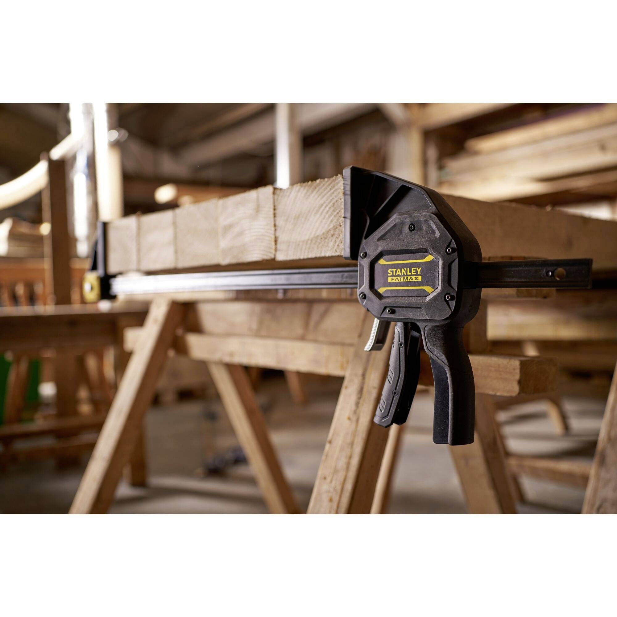 A workshop with a piece of wood on a workbench, secured by a Stanley clamp. The background shows blurred woodworking tools.
