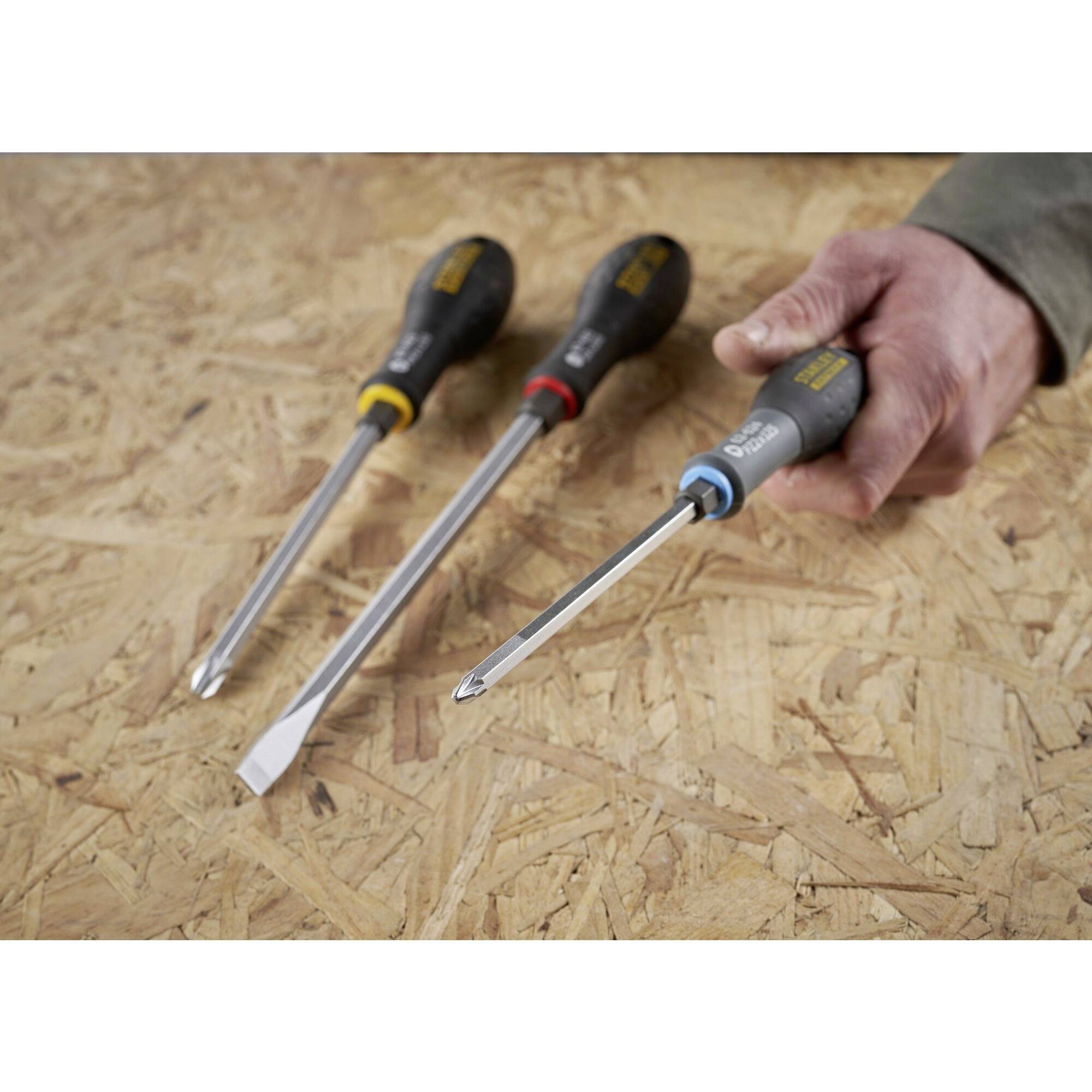 Three screwdrivers with differently coloured handles are lying on a wooden table. A hand is holding a screwdriver with a blue handle.