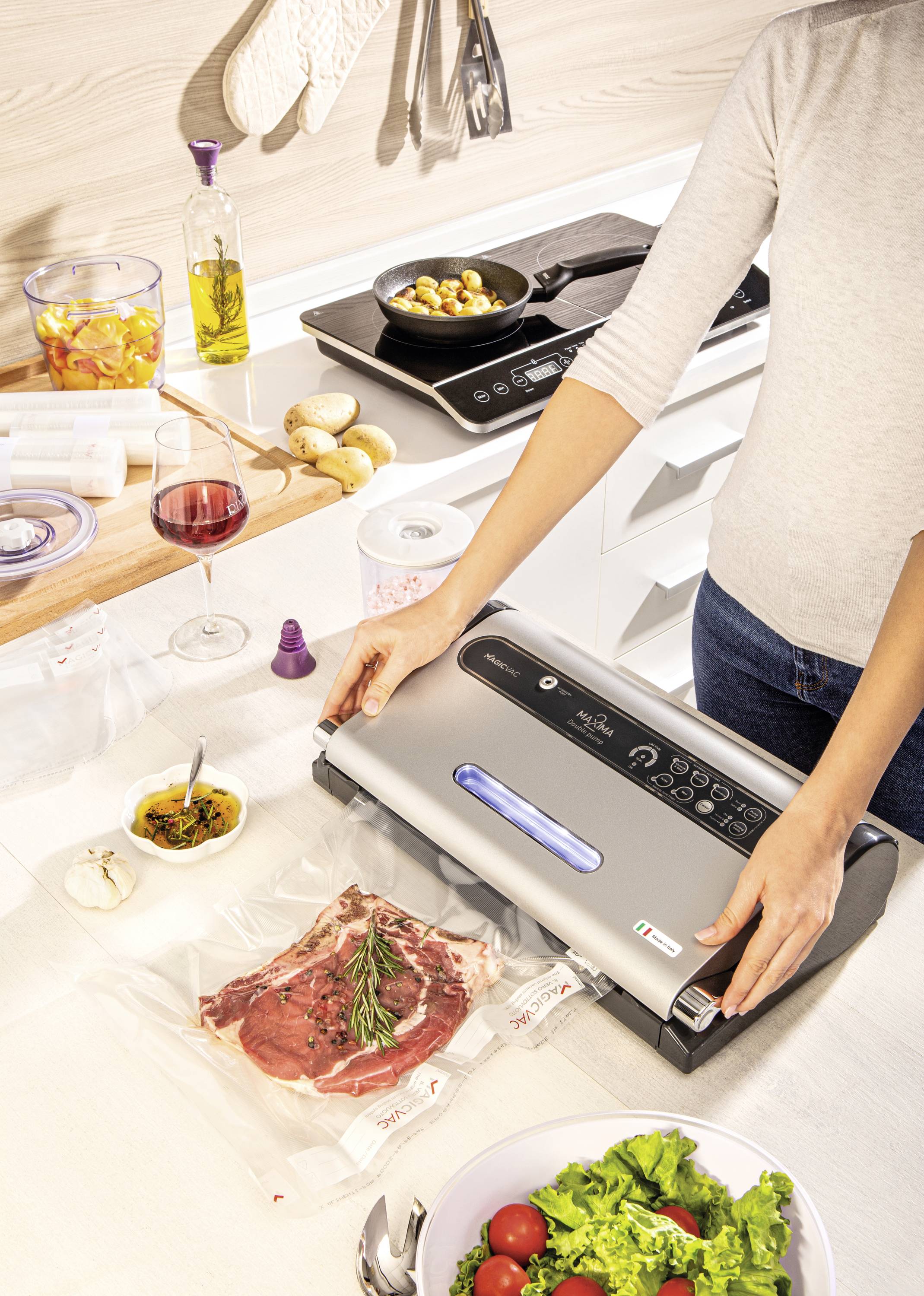 A person is using a vacuum sealing device in a modern kitchen to package a piece of meat with rosemary; cooking ingredients and utensils are visible in the background.