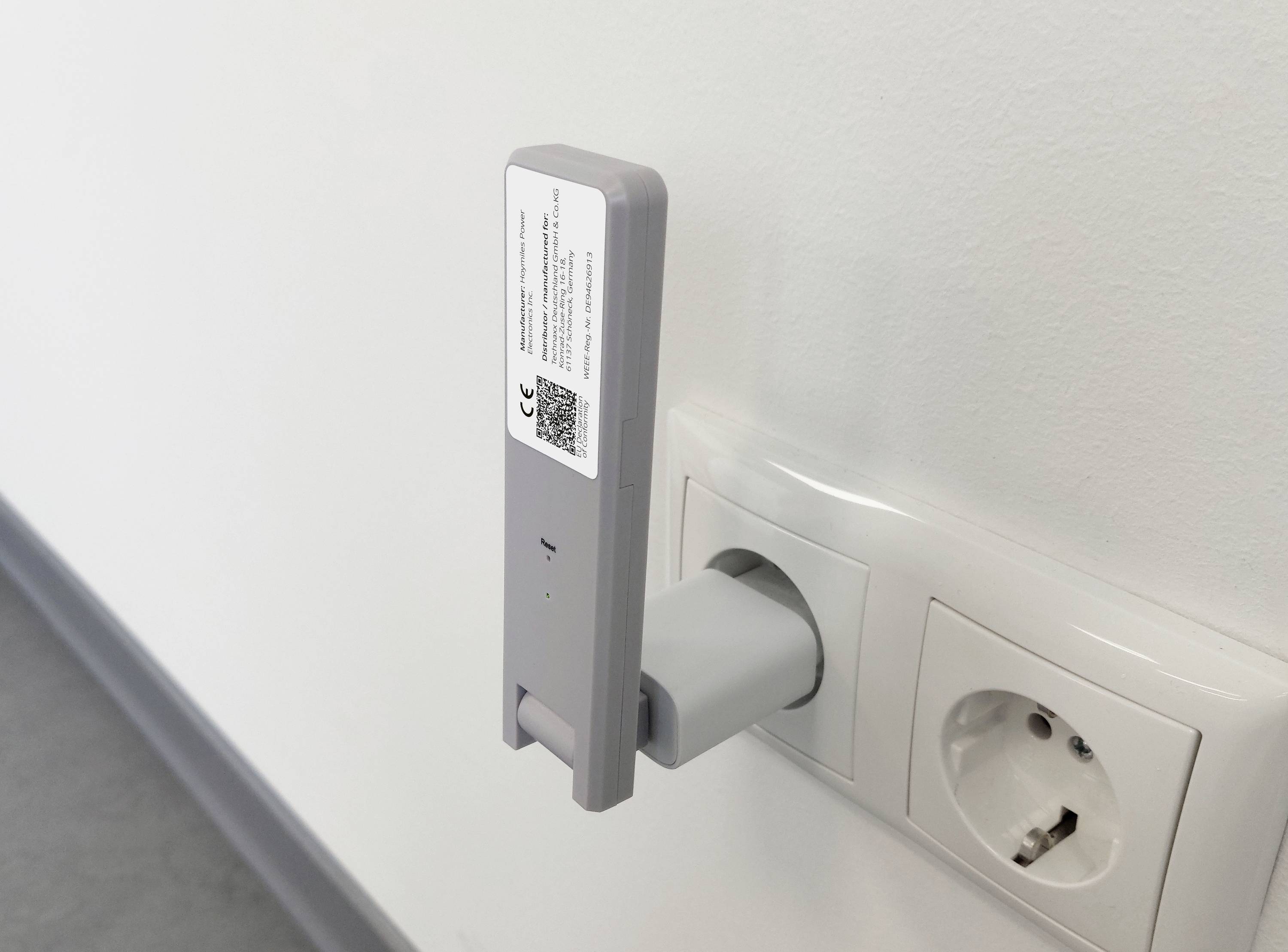 A grey device is plugged into a wall socket. It has a QR code and text on its side. Next to it is an empty socket.