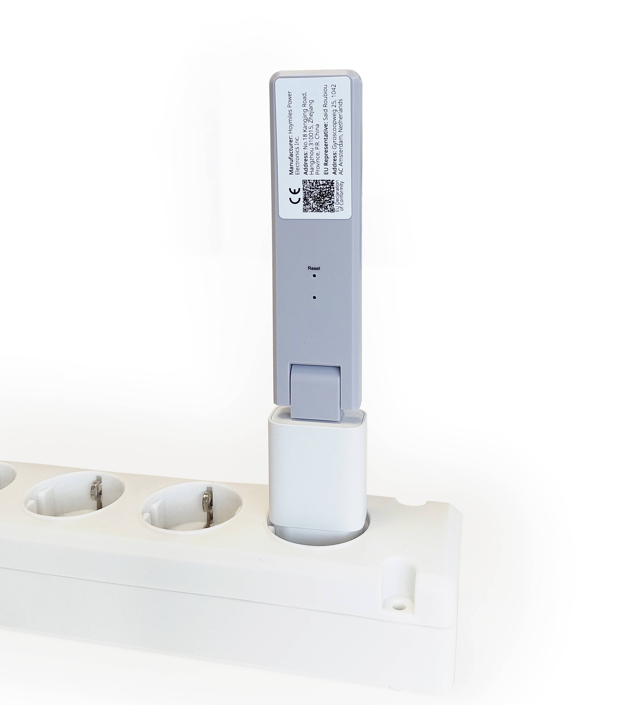 A grey device is plugged into a white power strip. A label with small text and a QR code is located on the device.