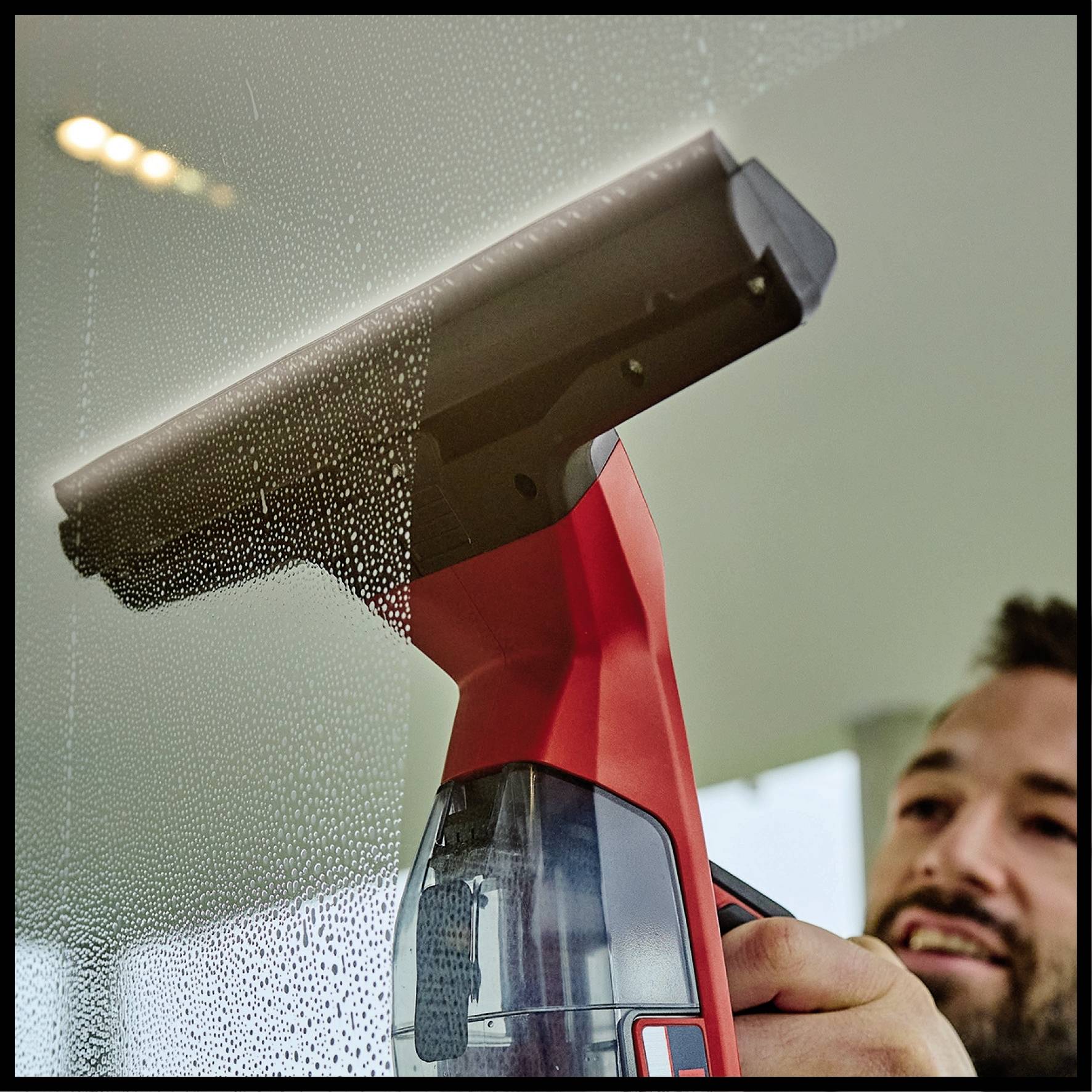 A person is cleaning a window with a red window squeegee, with lights blurred in the background.