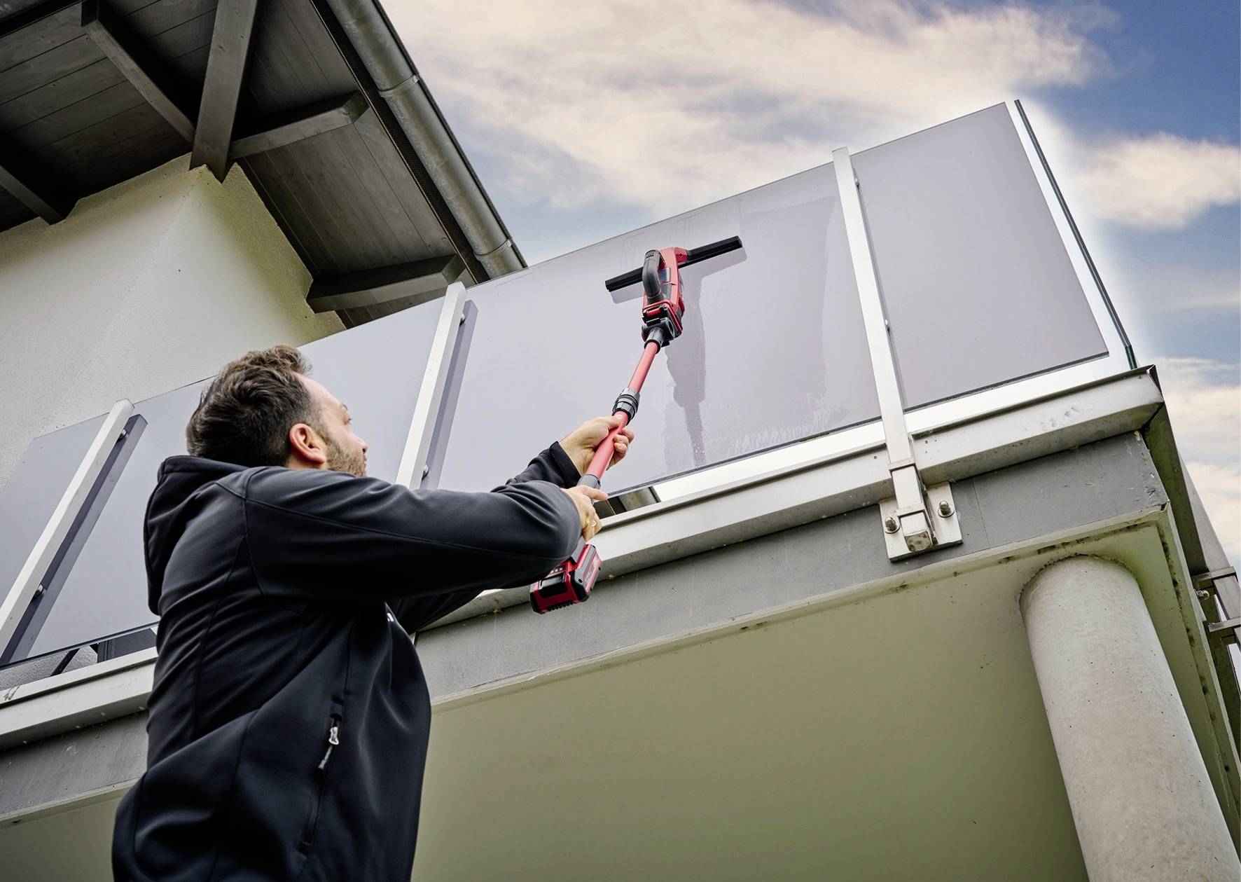 A person is cleaning the glass wall of a balcony from the outside using a long tool. The sky is partly cloudy.