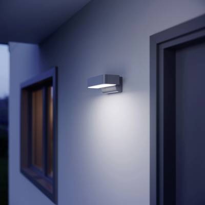 An external wall light illuminates a modern house entrance at dusk, with windows on the left and front door on the right visible.