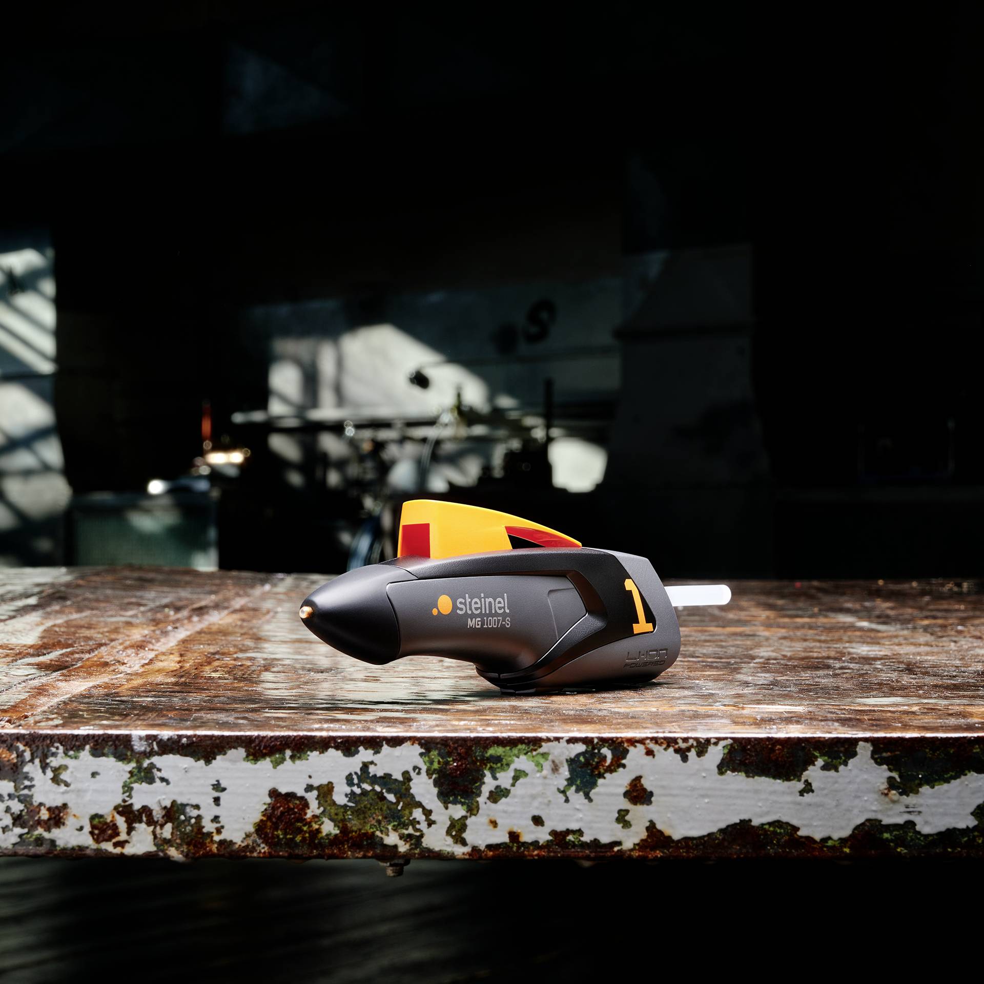A Steinel hot glue gun lies on a worn workbench in a workshop. Tools are blurred in the background.