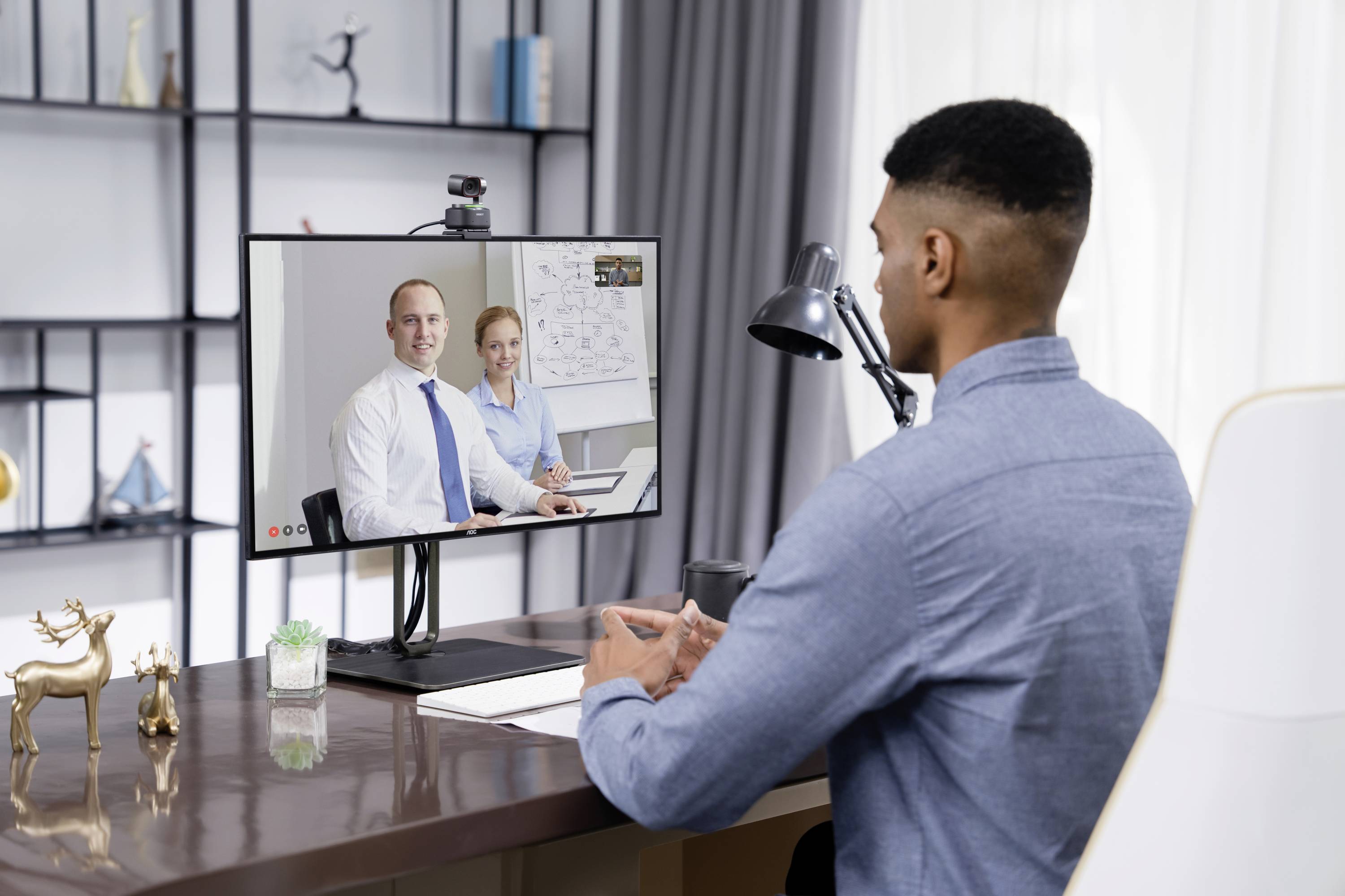 A man is sitting at a desk in a video conference with two colleagues on the screen. He has a laptop and notes in front of him.