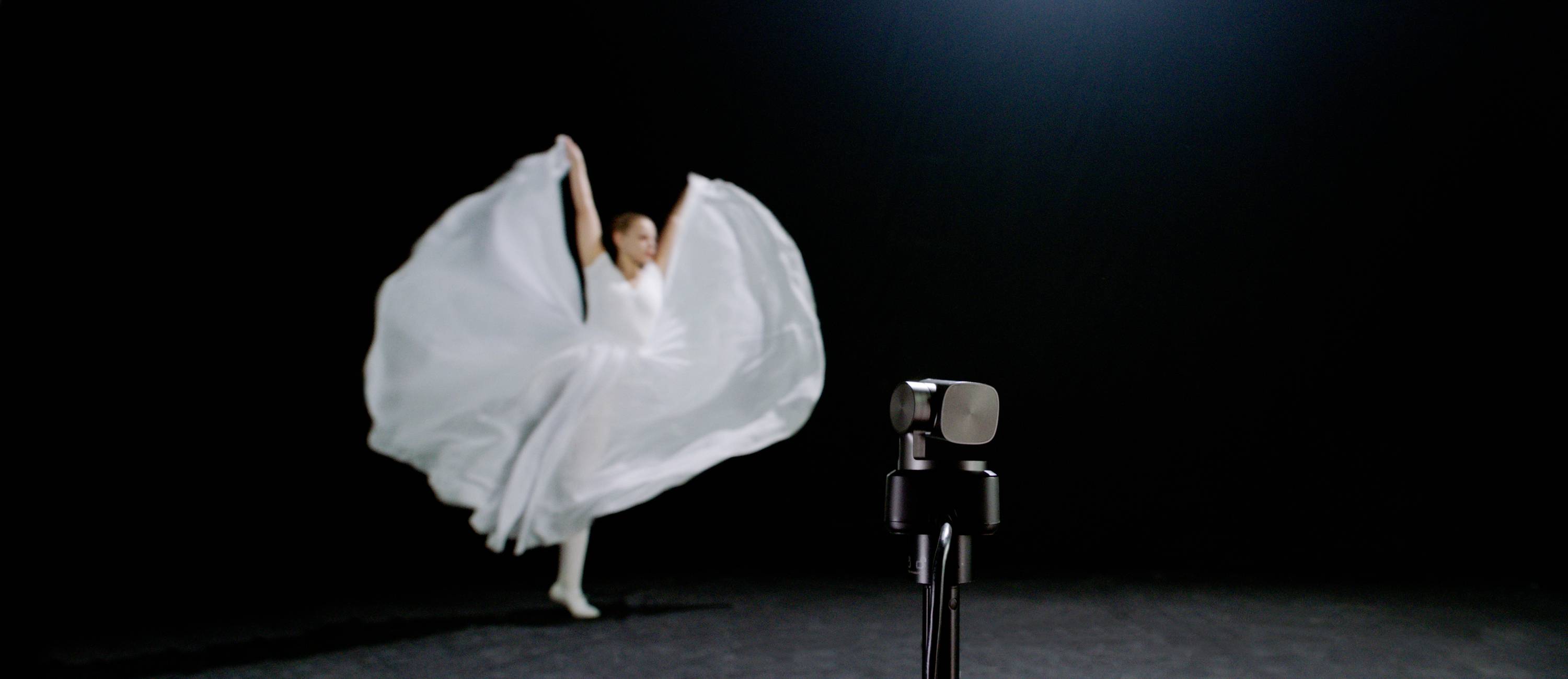 A dancer in a white dress twirls in the background, with a camera focused on her in the foreground, within a dimly lit room.