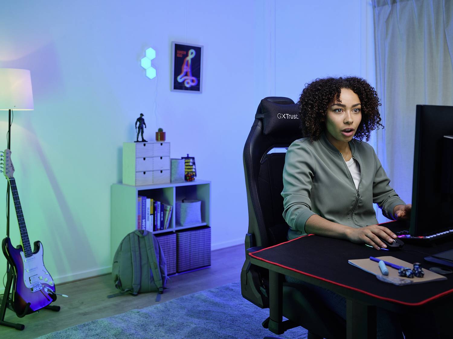 A person is sitting at a desk in front of a computer screen in a modernly furnished room with a guitar and shelving in the background.