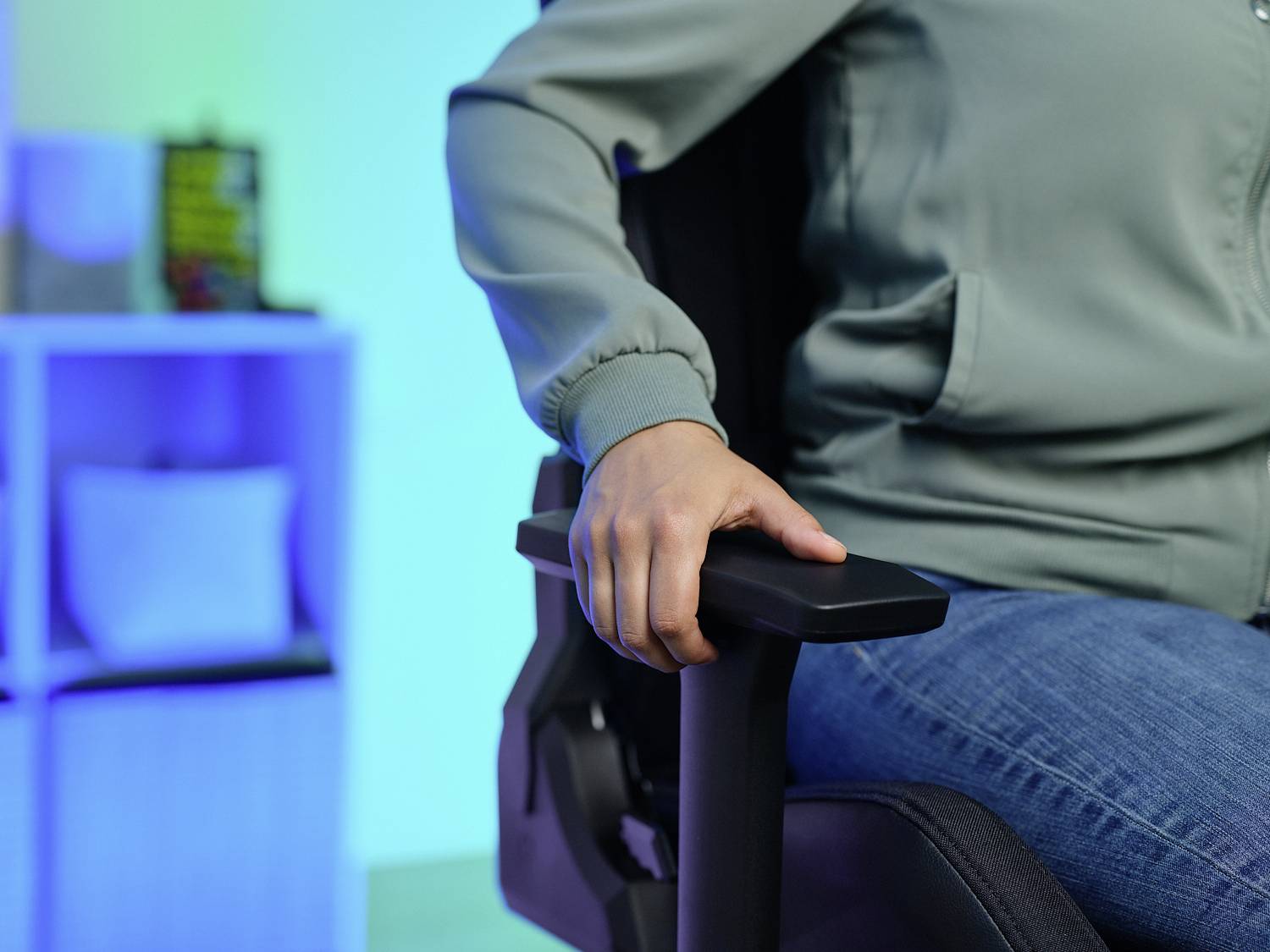 A person is sitting on an office chair, gripping the chair back. A bookshelf is blurred in the background.