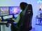 A person is sitting at a desk with two large monitors, playing a computer game. The room is illuminated with blue lighting.