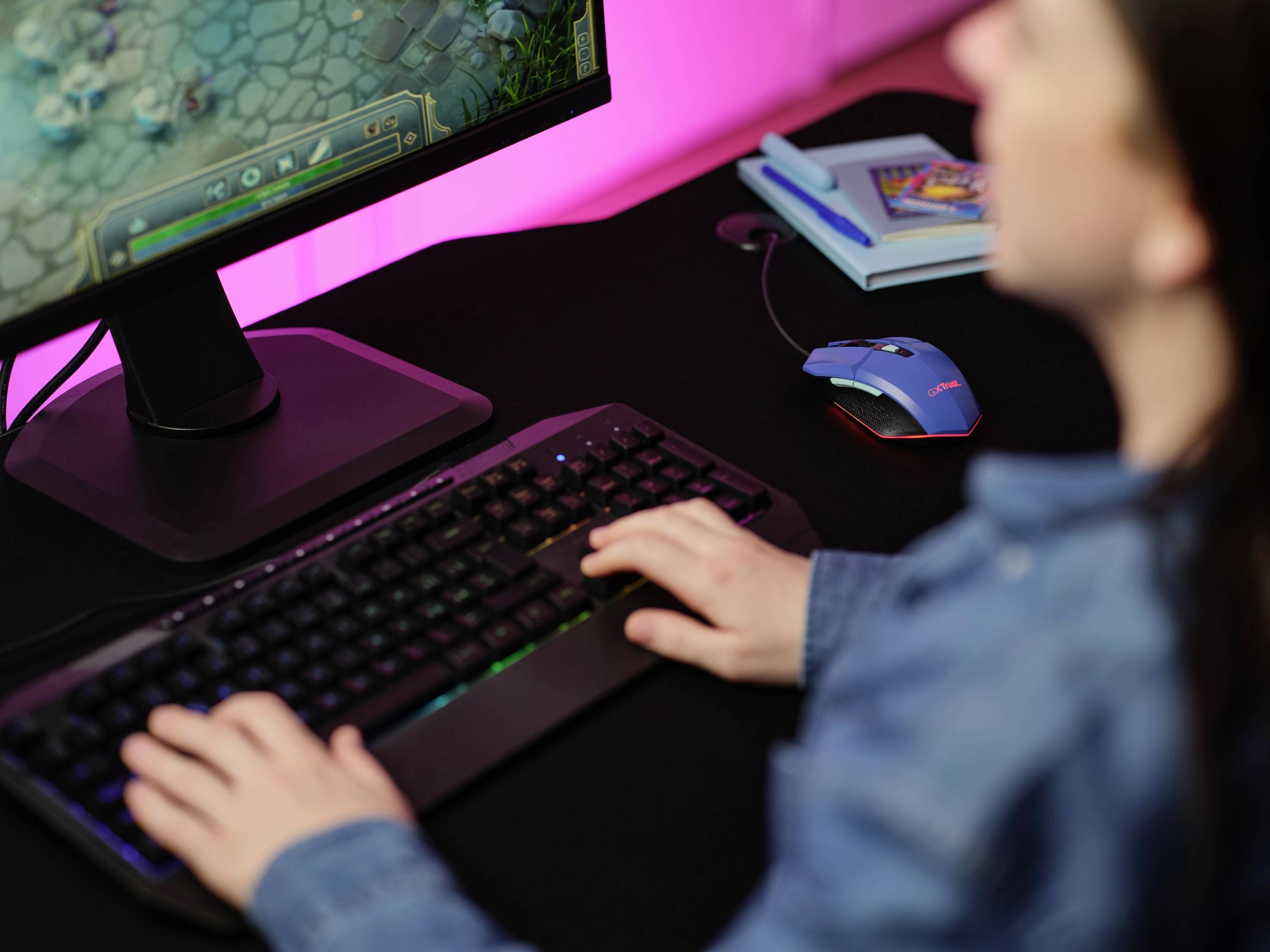 A person is playing a computer game on a monitor, with a keyboard and mouse on the desk. Notebooks can be seen in the background.