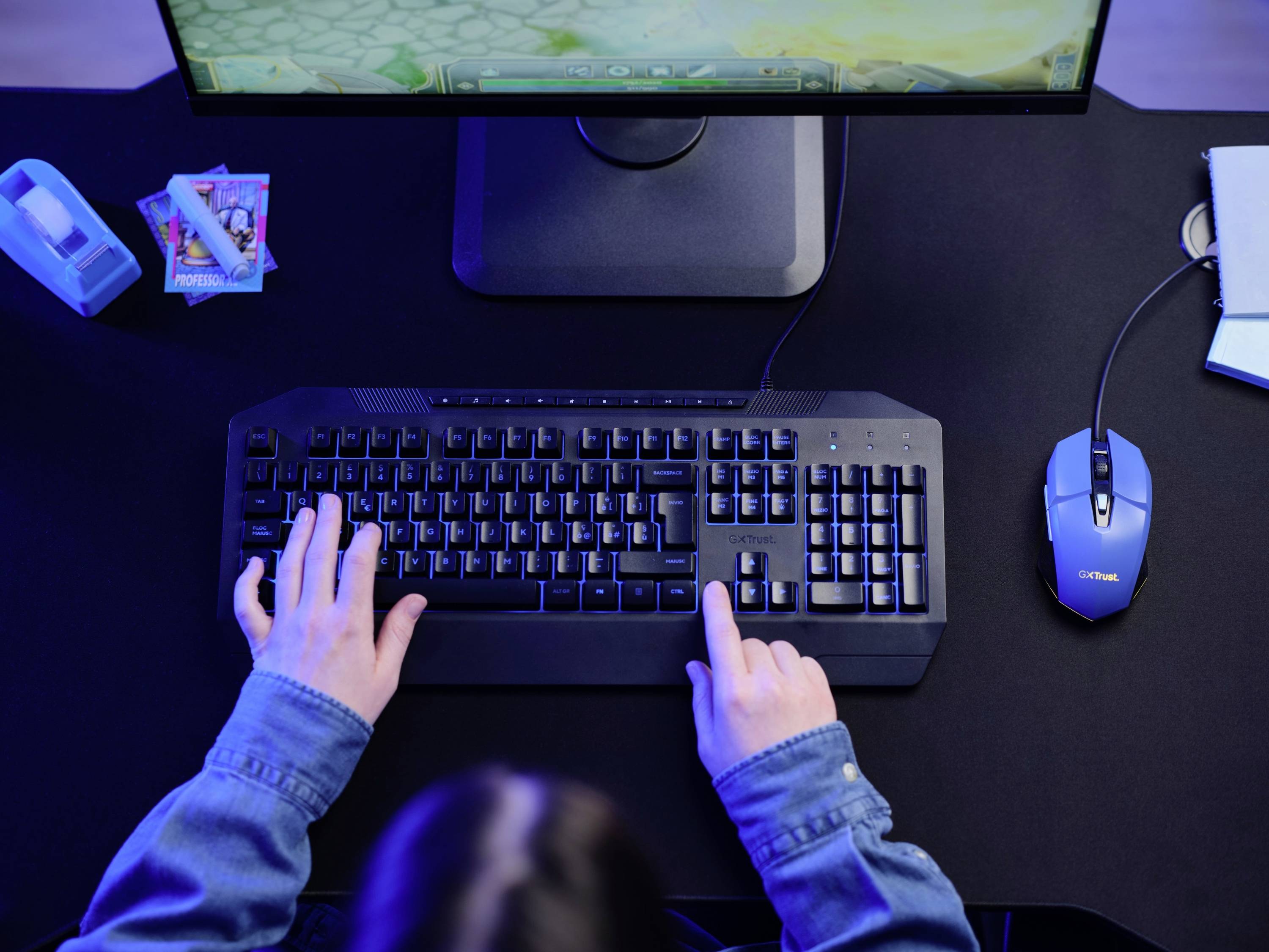 A person is typing on a keyboard and playing a computer game on a screen. A mouse is lying beside it, and cards are visible.