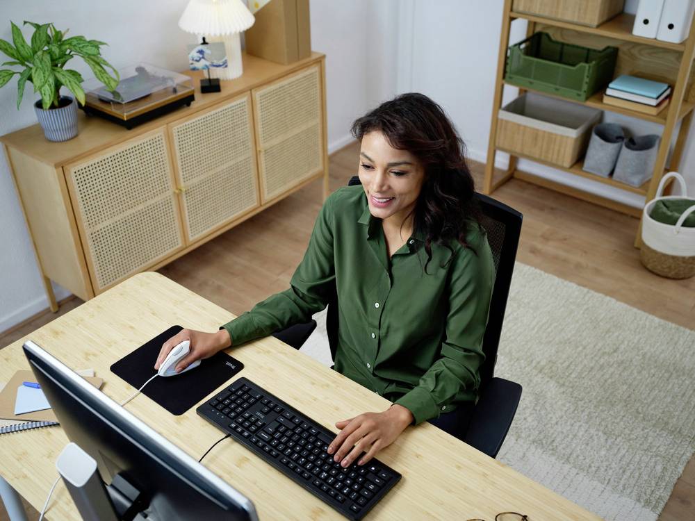 A woman in a green shirt is working at a desk with a computer. Shelves and plants are located in the background.