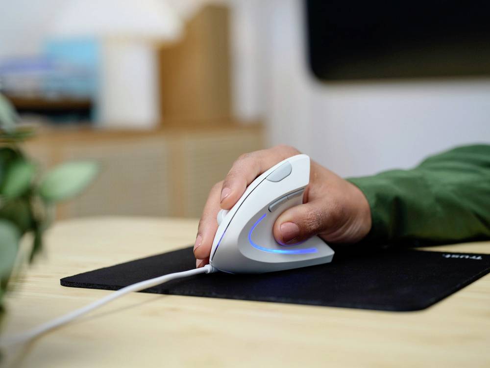 A person is using an ergonomic computer mouse on a desk. Blurred office objects can be seen in the background.
