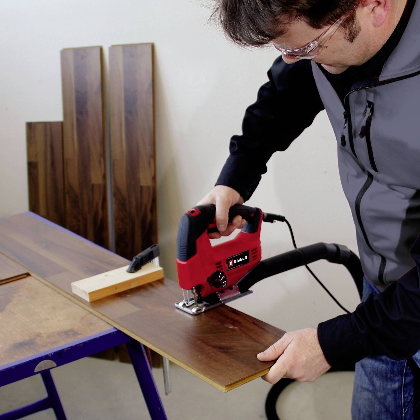 A person is cutting a laminate board with a handheld circular saw. Wooden boards are stacked against the wall in the background.