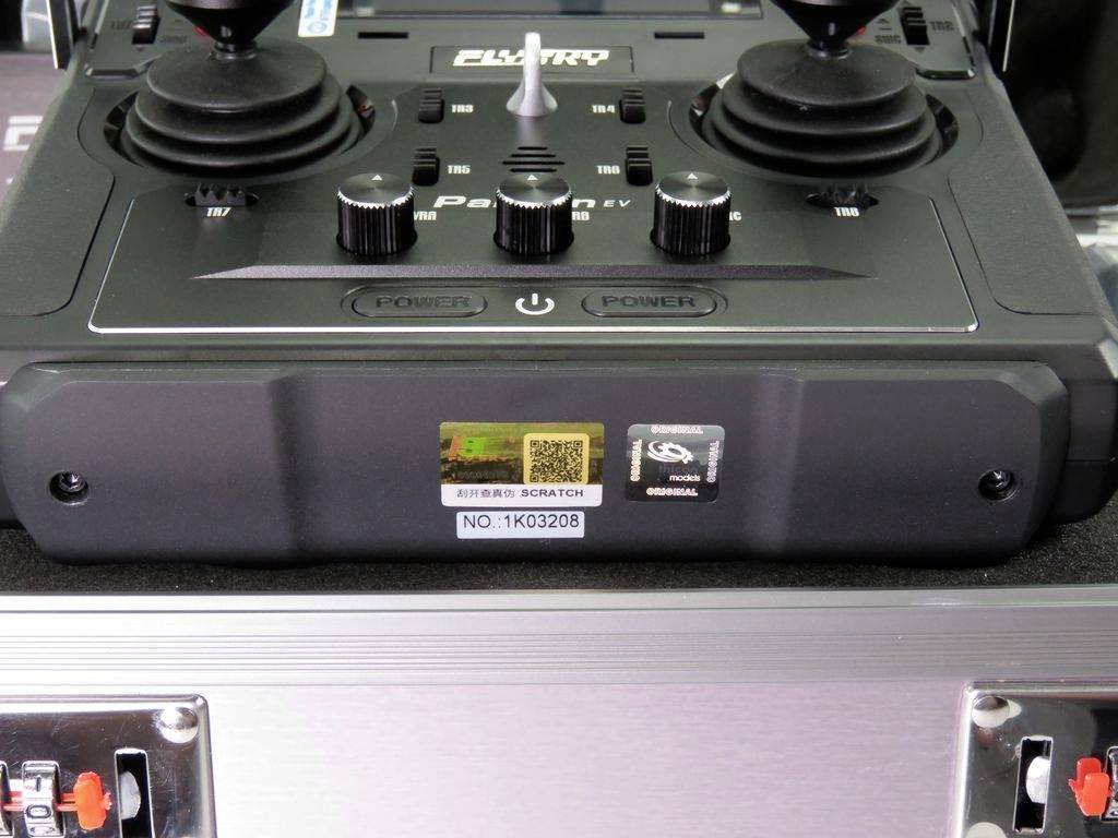 A black controller with two joysticks, buttons and a display, set against a metallic background. A label with a barcode is visible.