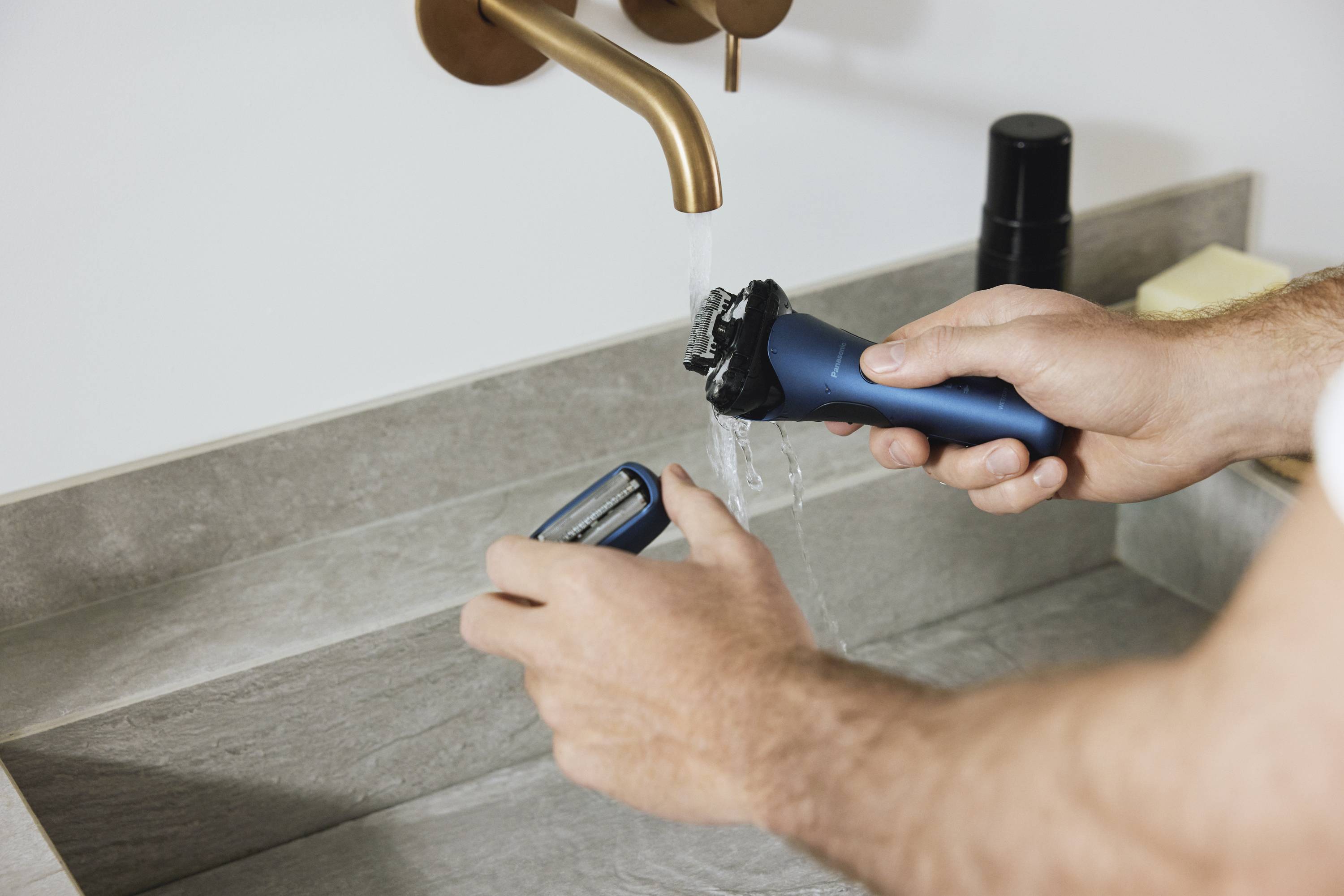 A person is rinsing an electric razor under running water. A tap and a shelf are visible.