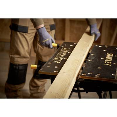 A person is cutting a long wooden board on a workbench. They are wearing work clothing and gloves. A workshop is visible in the background.