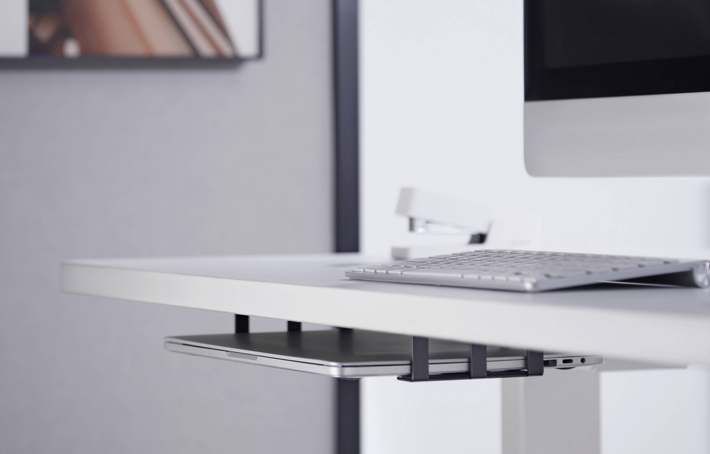 A modern desk with a keyboard and monitor arm. A laptop is stored in a shelf underneath. A blurred image is in the background.
