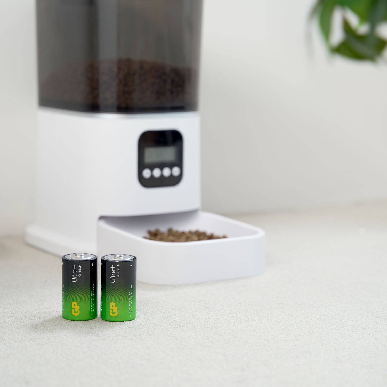 A feed dispenser drops dry food into a bowl. In the foreground are two GP batteries.