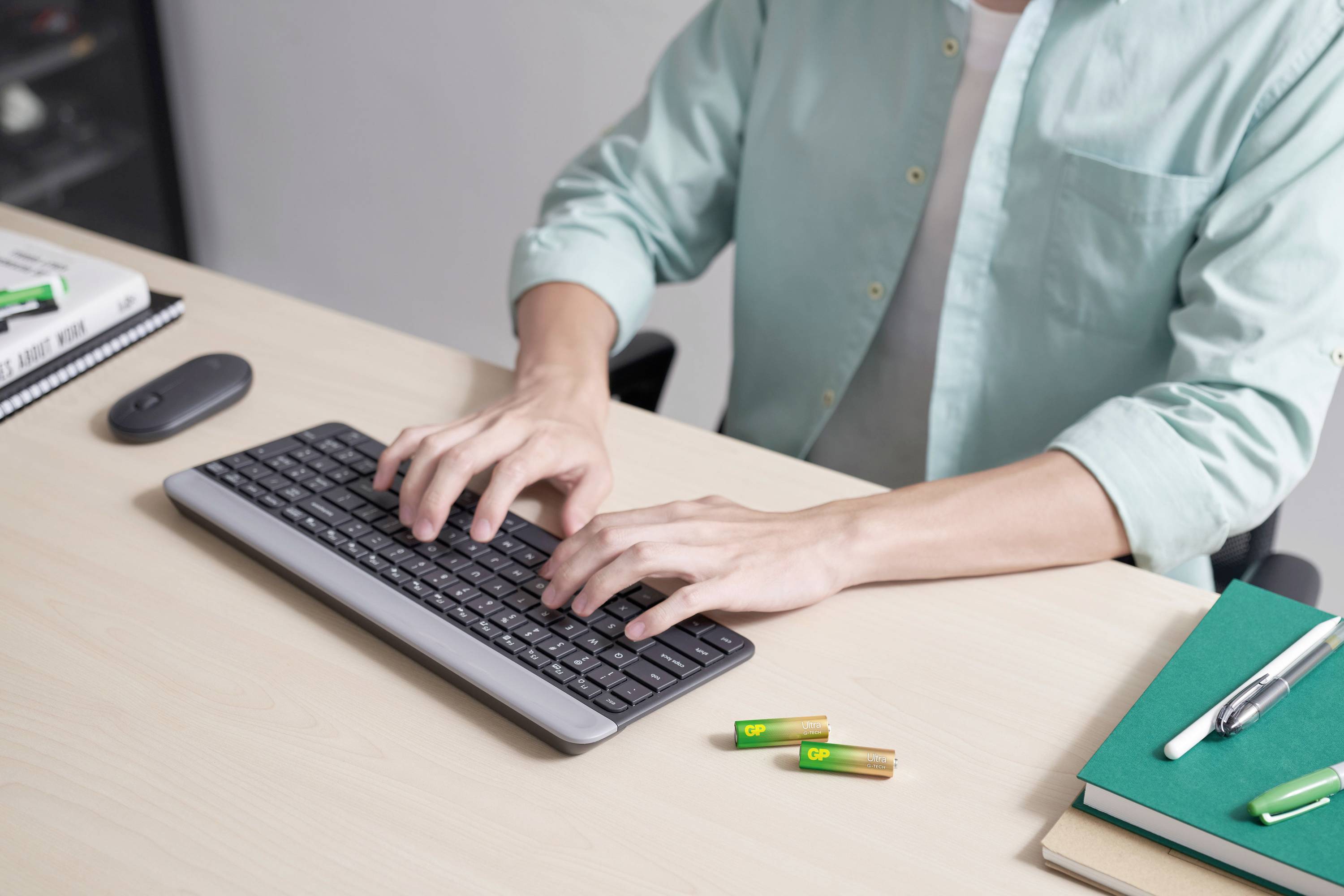 A person wearing green clothing is typing on a keyboard at a desk. Batteries, a book, and a pen are lying on the desk.