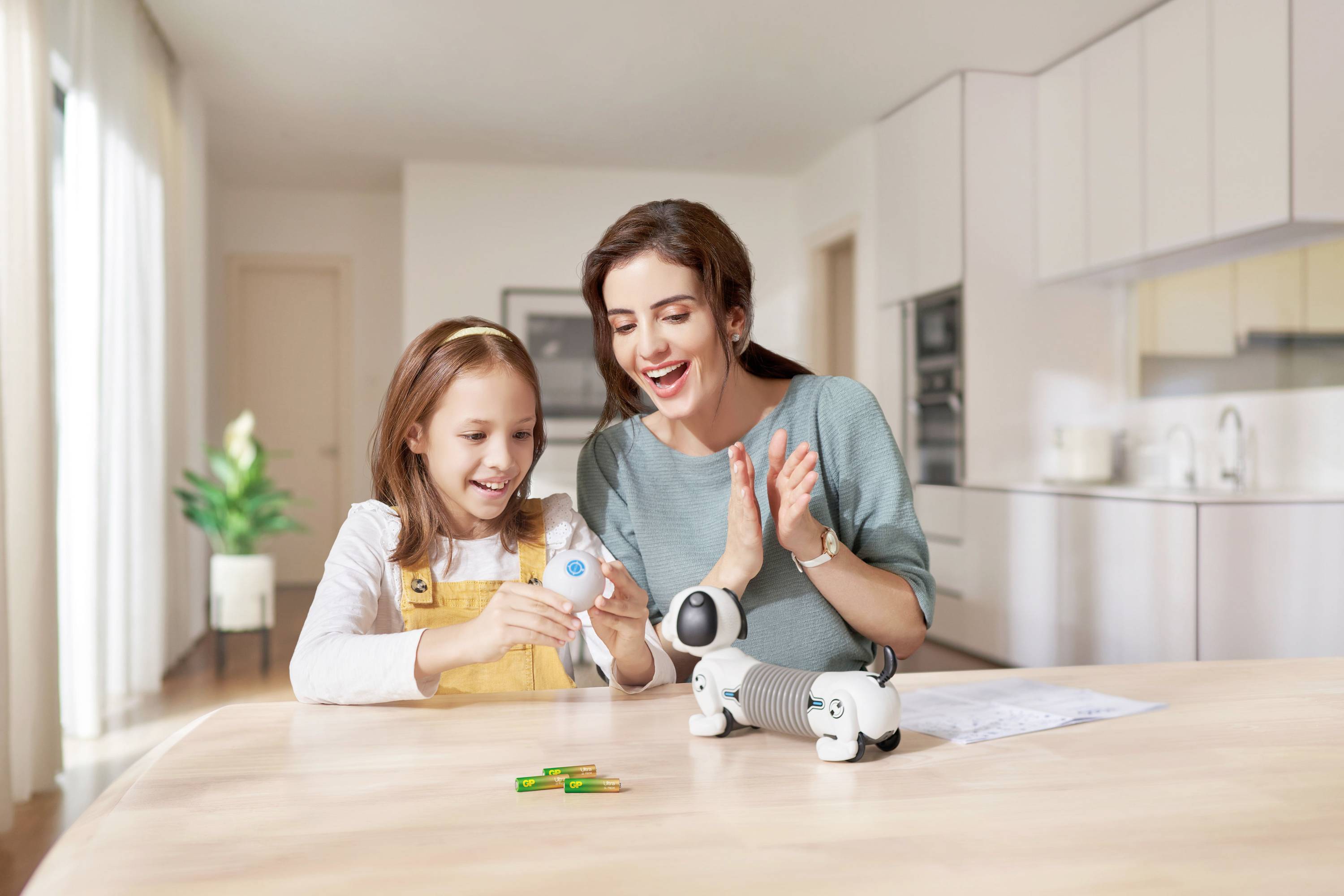 Mother and daughter at the table, interacting with a toy robot in a bright living room. Both look cheerful.