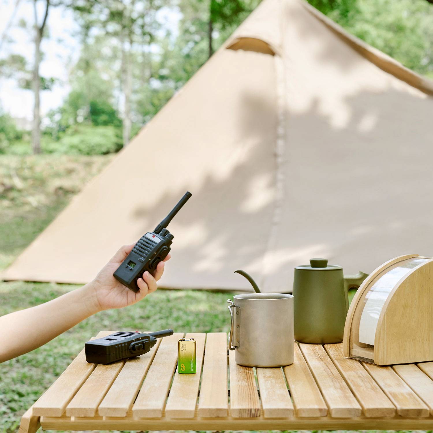 A table with a radio, mug, and bread bin in front of a tent outdoors. Atmospheric scene of a campsite.