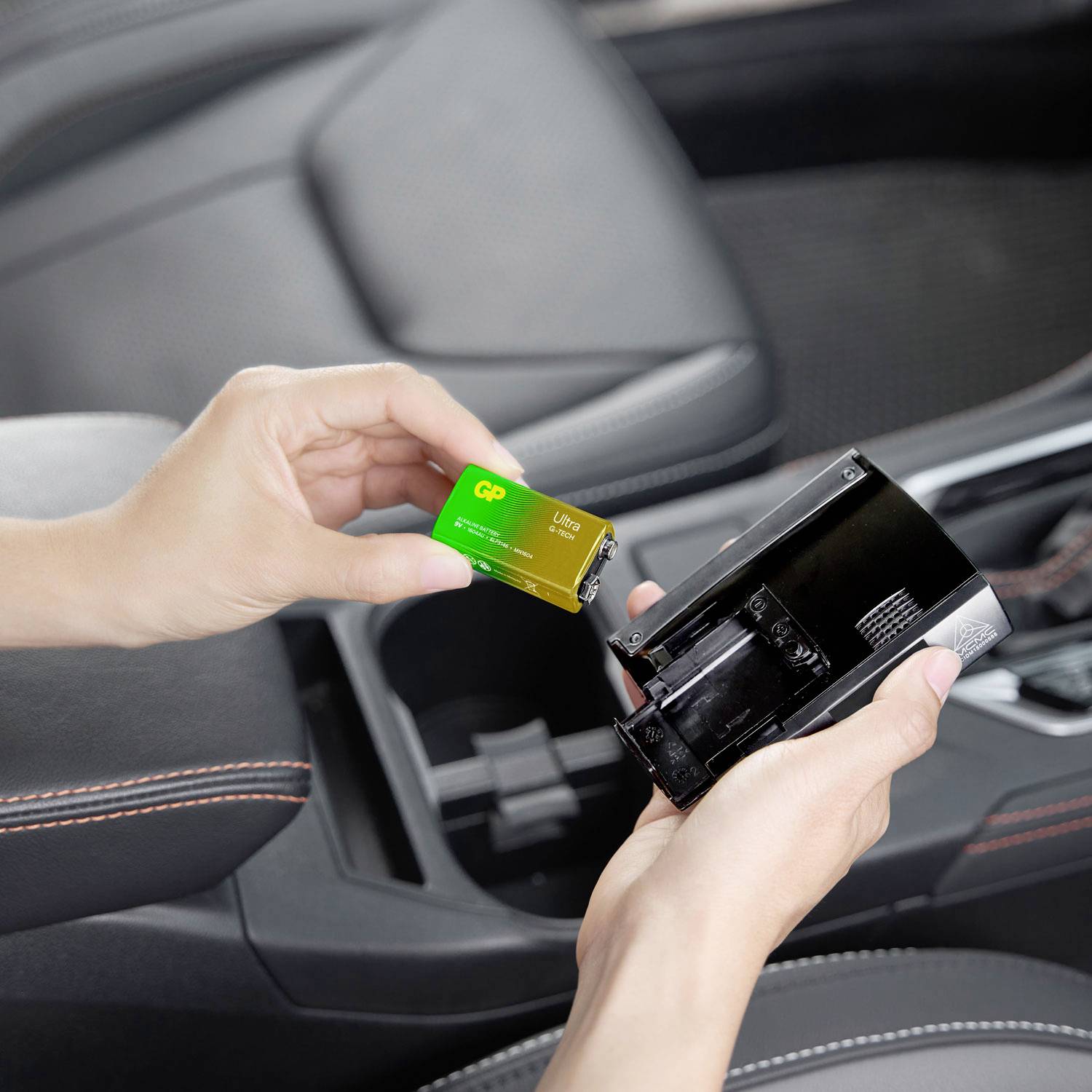 A person is holding a green 9V battery and inserting it into a compact black charger positioned in a car.