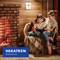 A woman and a child sit by a lit fireplace, reading together. A fire safety device is mounted on the wall. The brand logo, 'HEKATRON Brandschutz,' is visible.