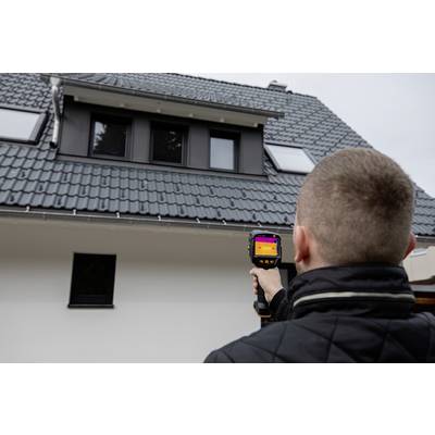 A person is pointing a thermal imaging camera at the roof of a house. The camera displays a thermal image to check the insulation.