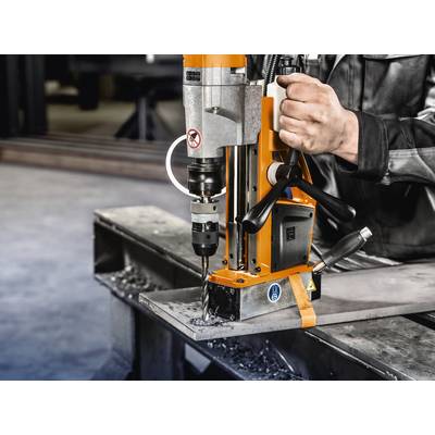 A person is operating an orange magnetic drilling machine on a metal beam. Metal shavings are visible around the drilling site.