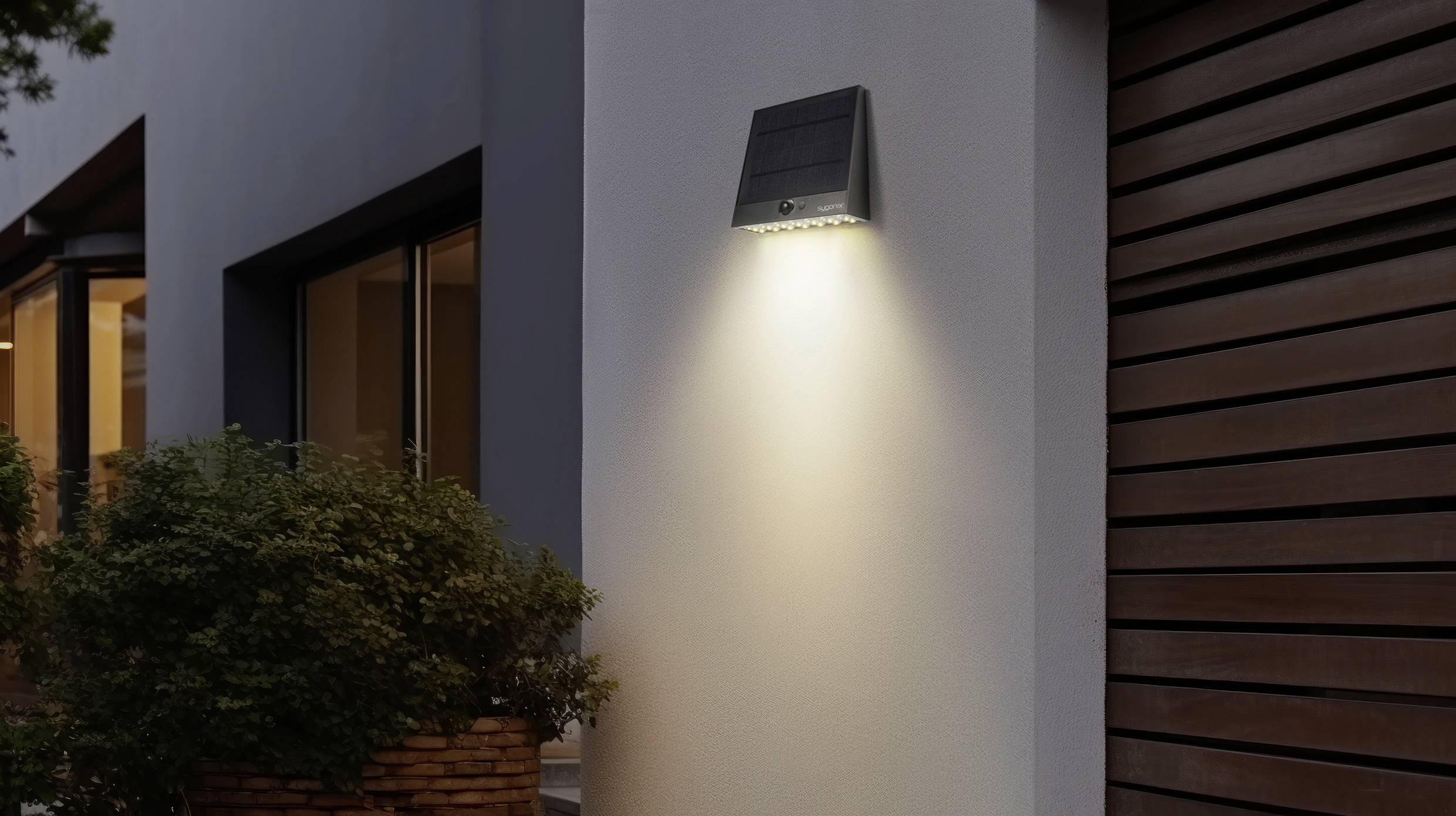 Modern exterior wall with a switched-on solar lamp illuminating the area beneath it. Windows and green plants are located to the left.