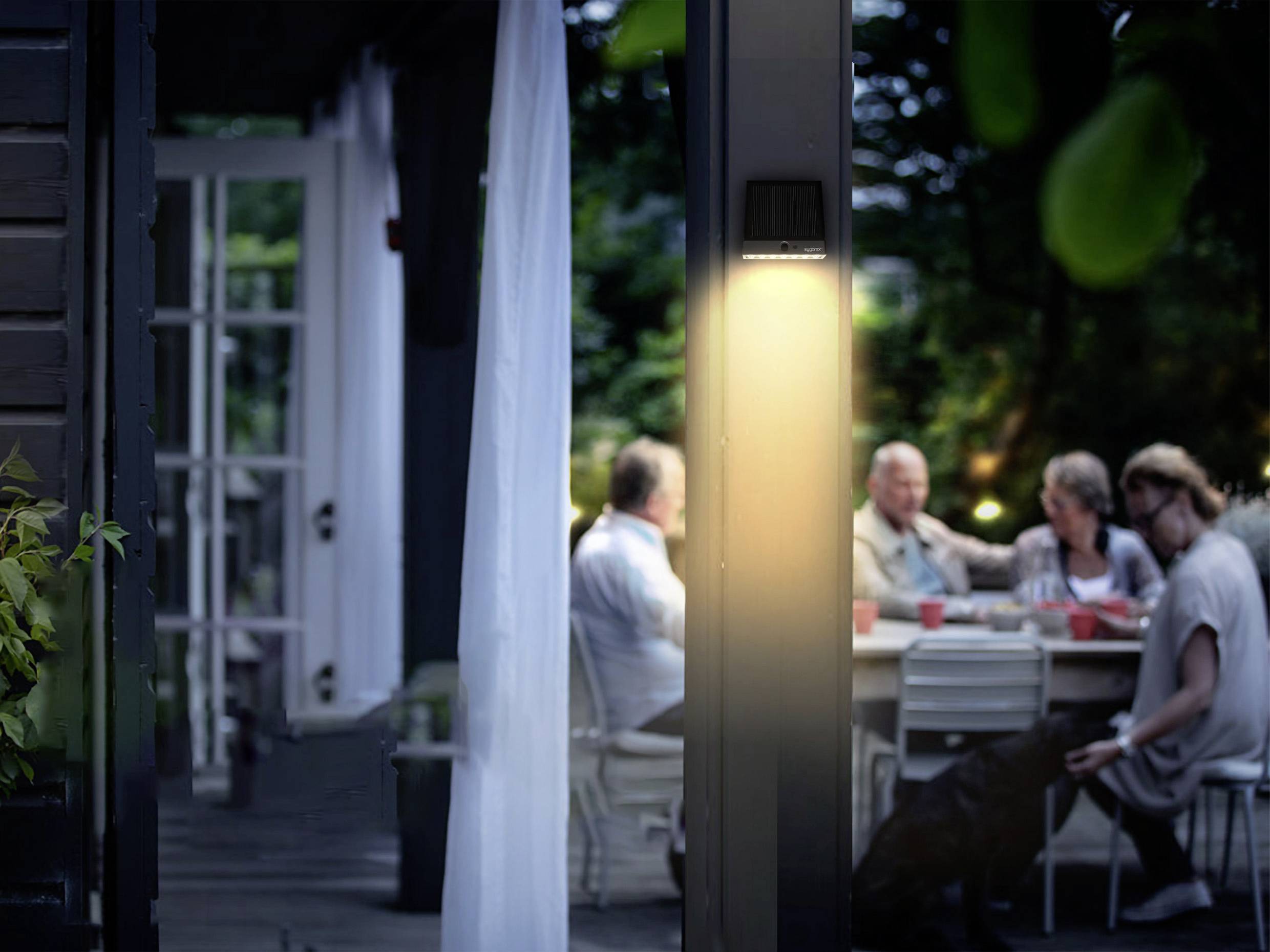 A group of people sit around a table in the garden in the evening, illuminated by a warm lamp. A relaxed conversational atmosphere prevails.
