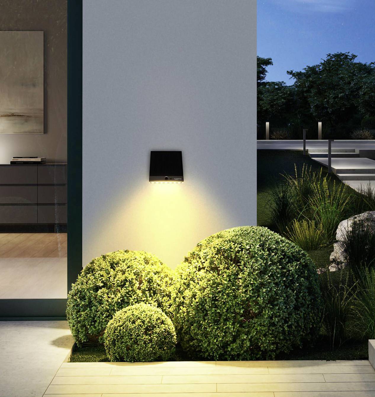 An outdoor wall light illuminates three green box trees in front of a modern building in the evening, creating a warm atmosphere.