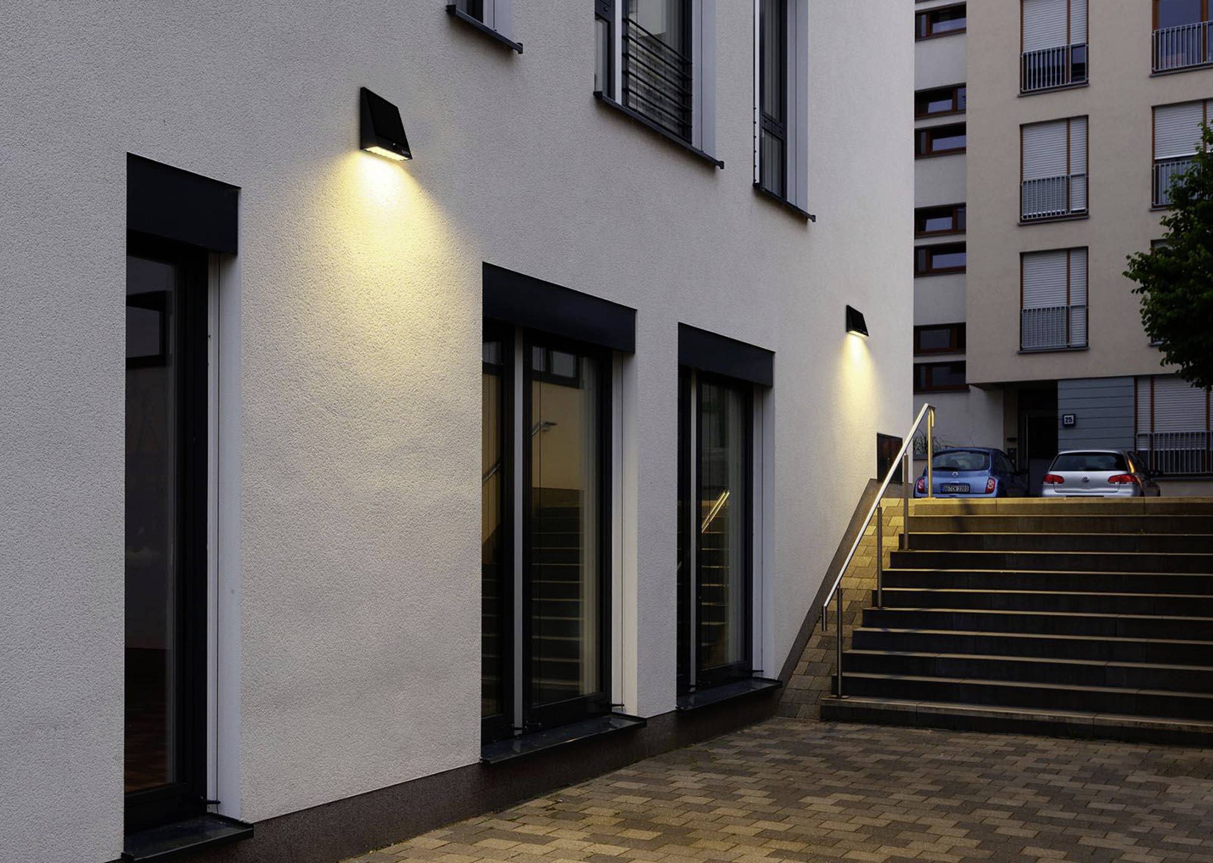 A modern building view with evening illumination on the facade, with a staircase on the right leading to parked cars in the background.